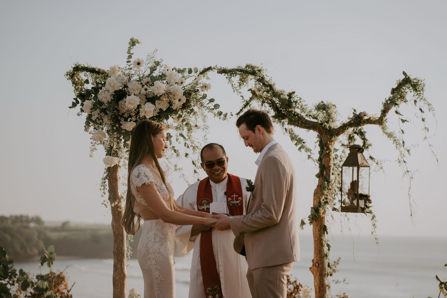 A couple exchanges vows during a romantic lakeside wedding ceremony under a rustic wooden arch decorated with flowers.