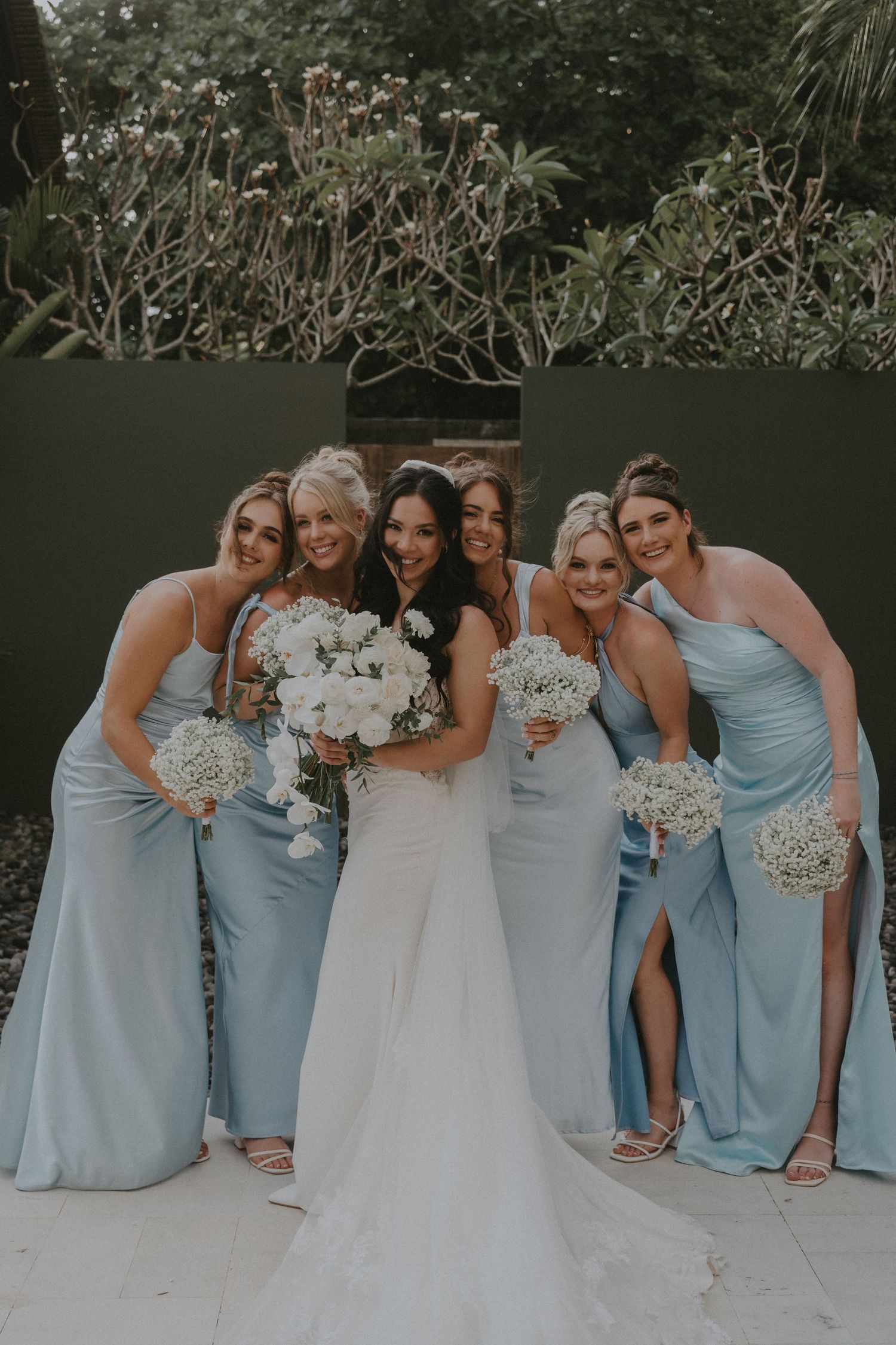 Bridesmaids in light blue dresses pose playfully with bride in white gown holding bouquets during outdoor wedding photos.