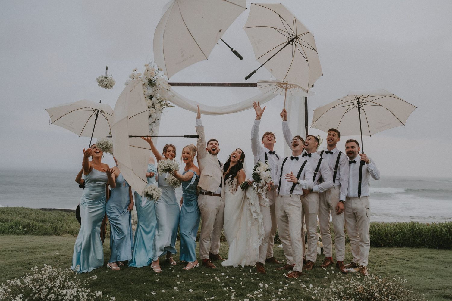 Wedding party in turquoise dresses and khaki suits pose with white umbrellas on a coastal cliff overlooking the ocean.