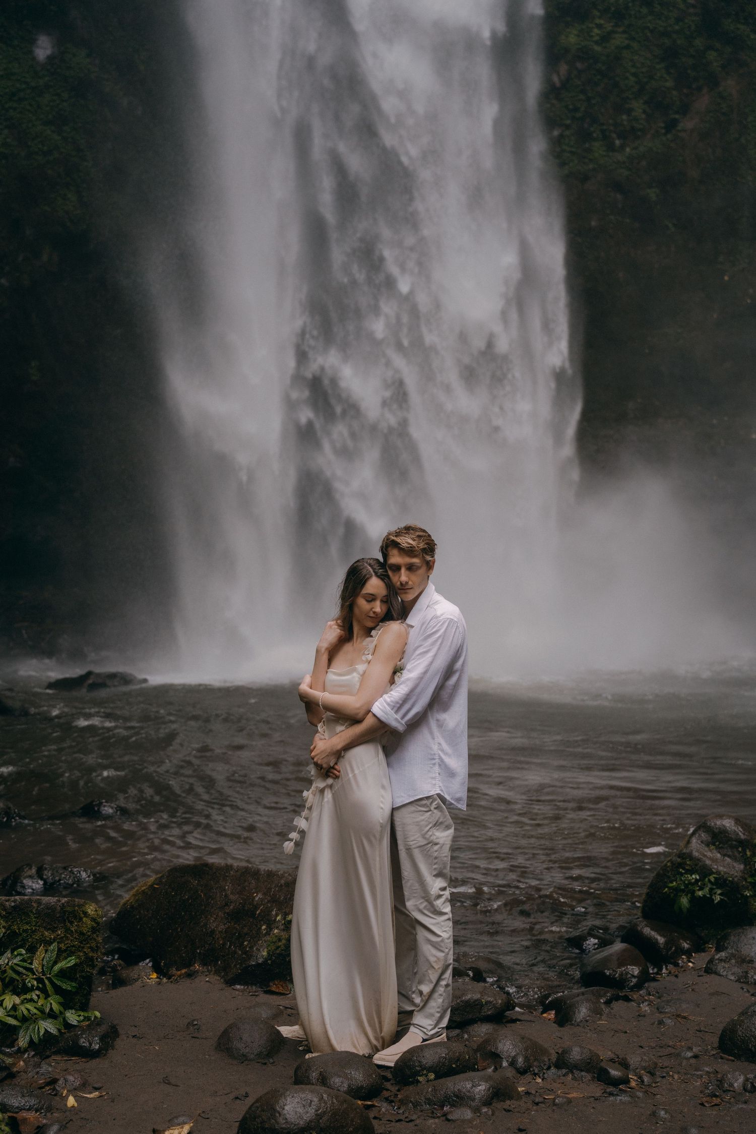 A romantic couple embraces near a misty waterfall against a dramatic natural backdrop.