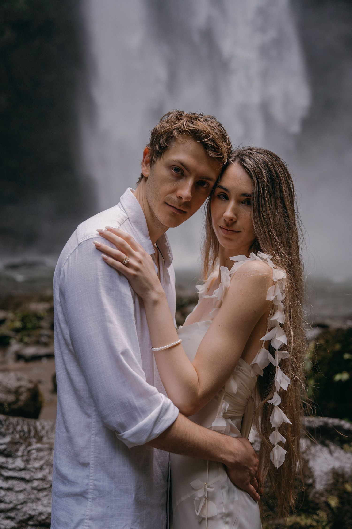 A romantic couple embraces near a misty waterfall, with the woman wearing a white dress and the man in a light colored shirt.