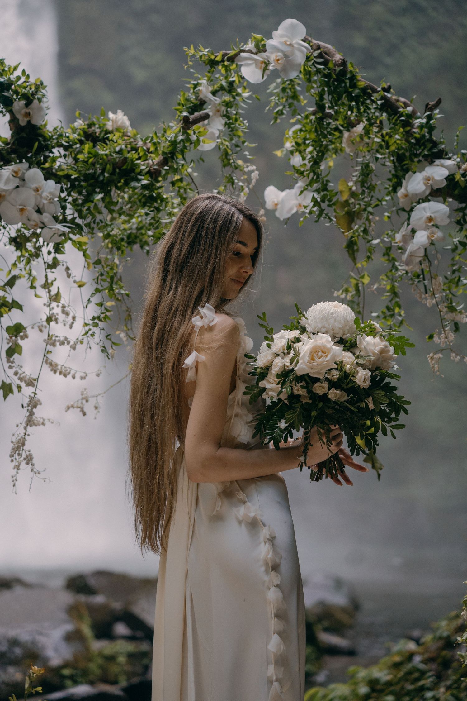 A romantic garden scene with white orchids and roses framing a flowing cream dress against misty natural backdrop.