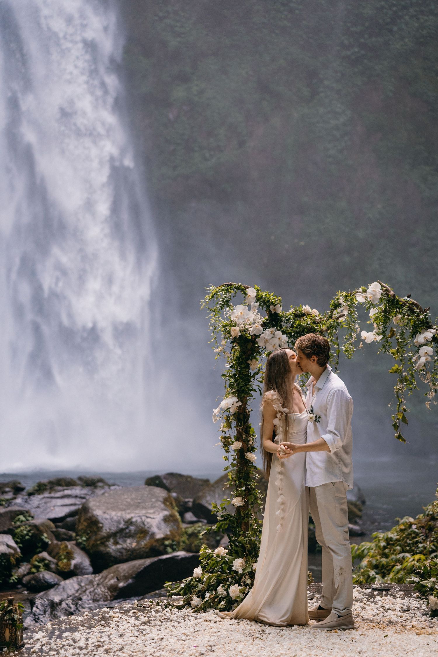 A romantic moment unfolds by a majestic waterfall with a floral arch creating an intimate wedding ceremony setting.