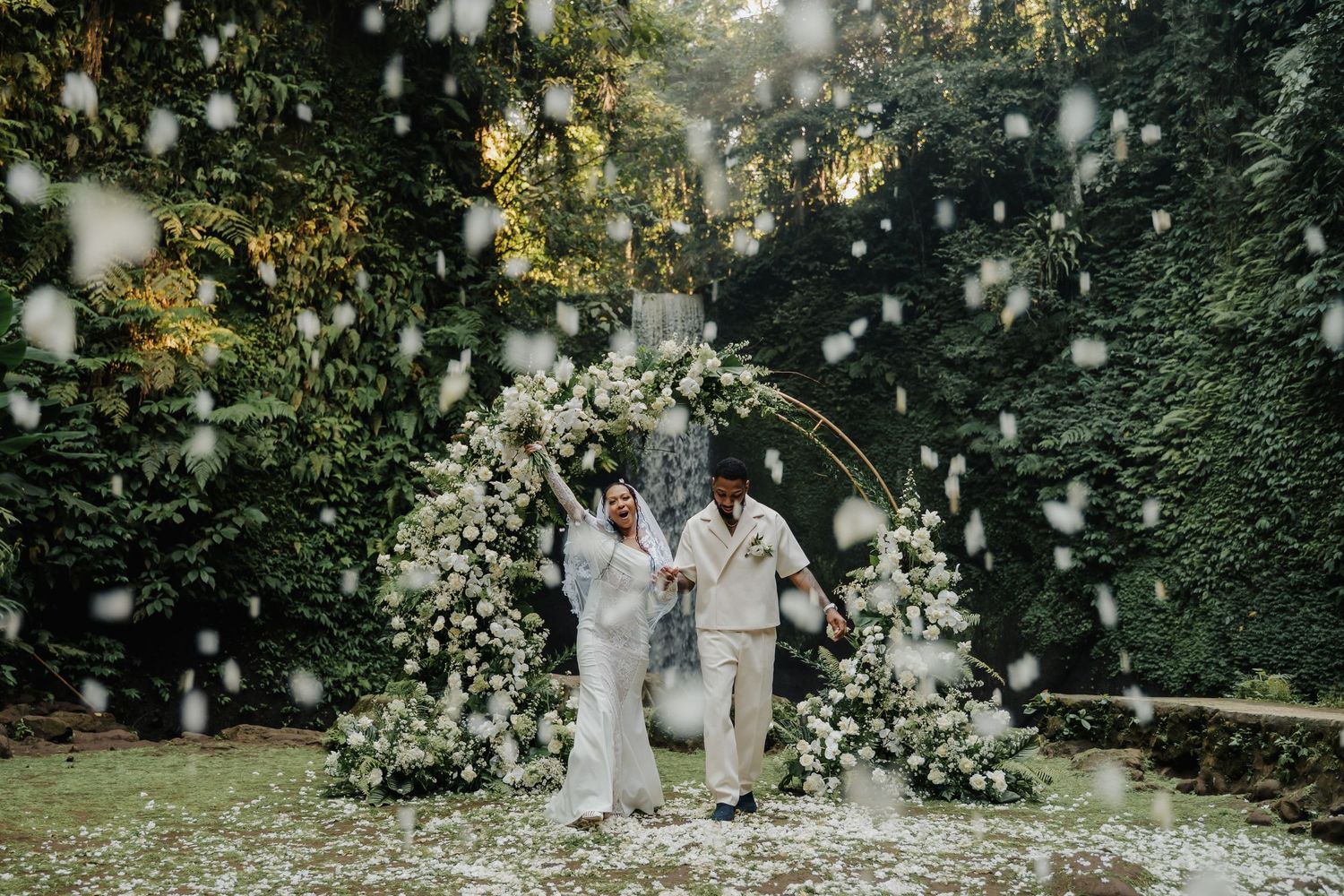 Newlyweds walking through shower of white flower petals in front of lush green floral wedding arch.