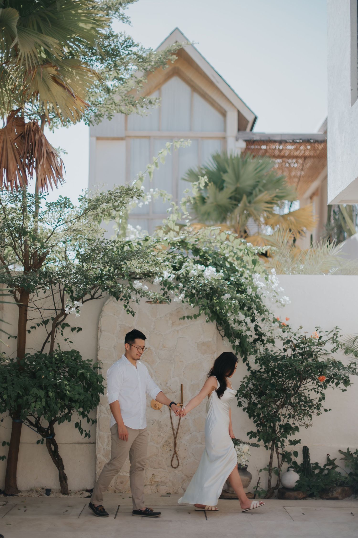 Couple holding hands walking near a white floral arch against a rustic wall with lush greenery and a modern building backdrop.