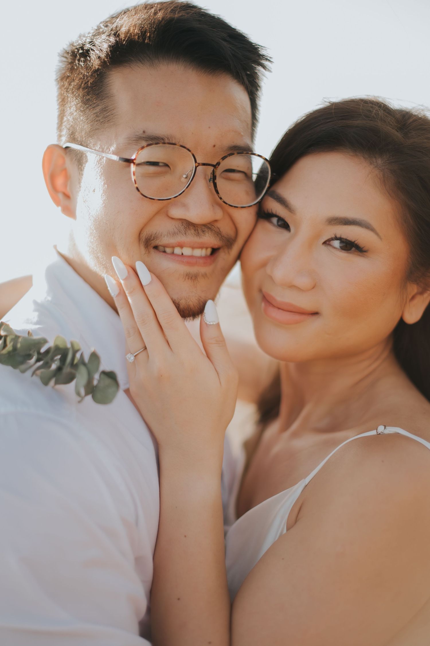 Happy couple posing for engagement photo, woman showing diamond ring, man wearing glasses, outdoors in soft natural light.