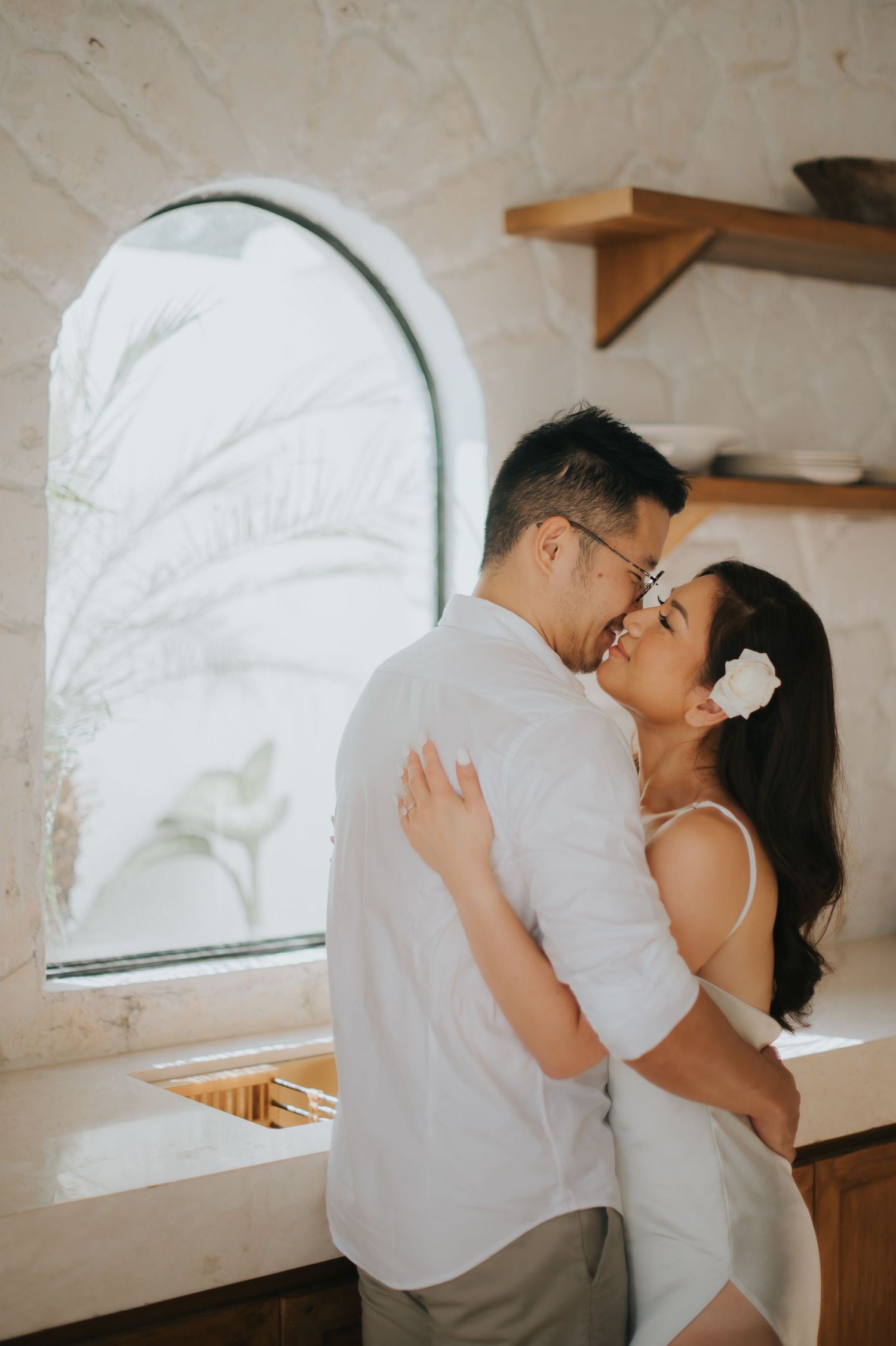 Couple embracing and smiling near an arched window in a bright white interior, woman wearing a white flower in her hair.