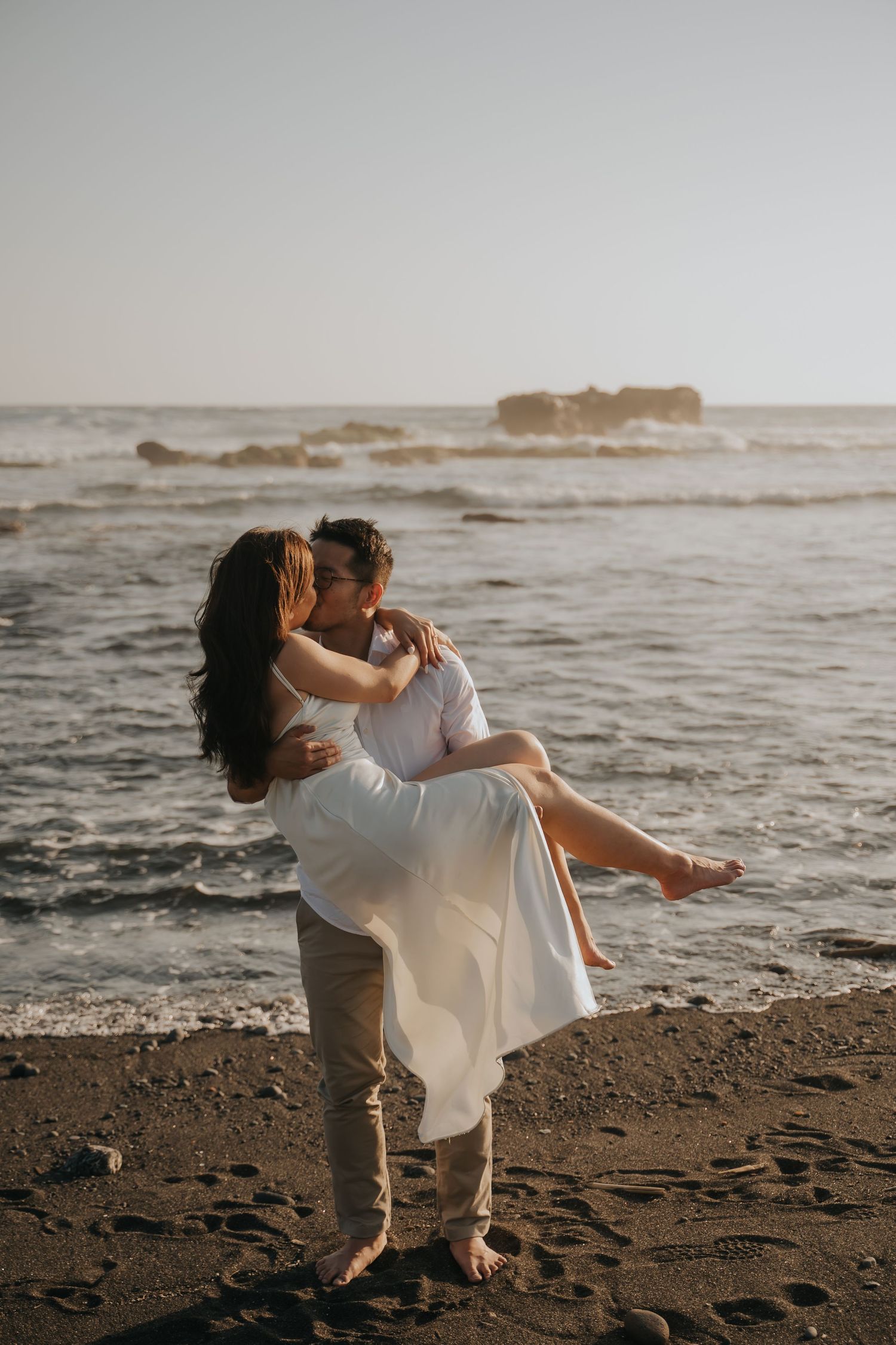 Couple in white embracing on a dark sandy beach, man lifting woman with ocean waves in the background.