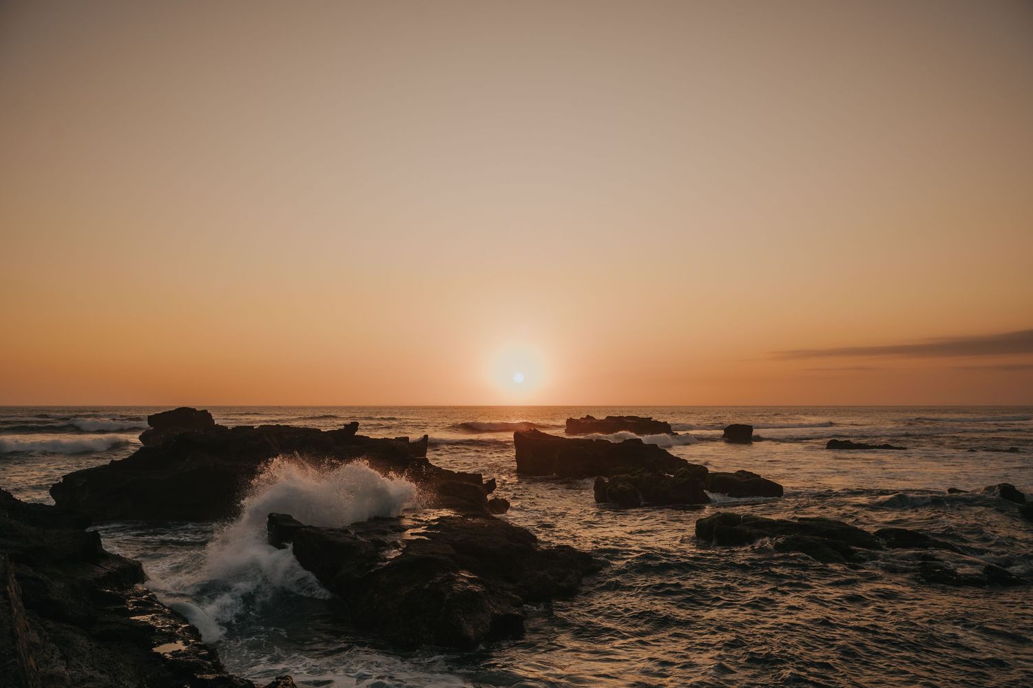Waves crash against rocky shore at golden sunset over ocean horizon.