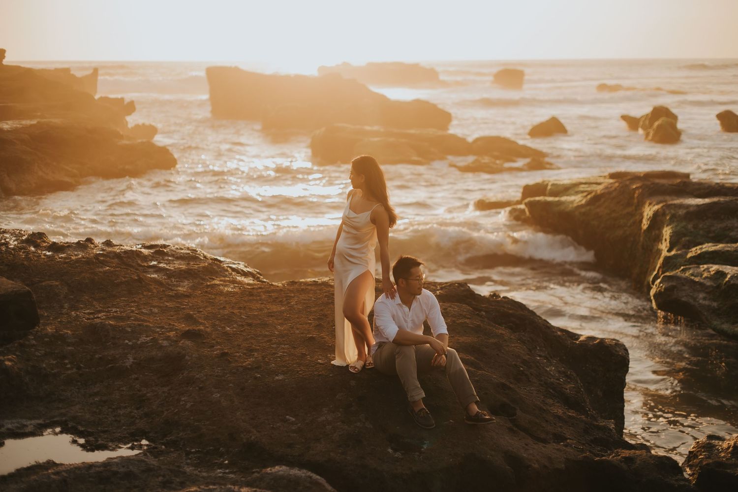 Couple posing on rocky coastline at golden sunset, waves crashing behind them in a romantic engagement photo.