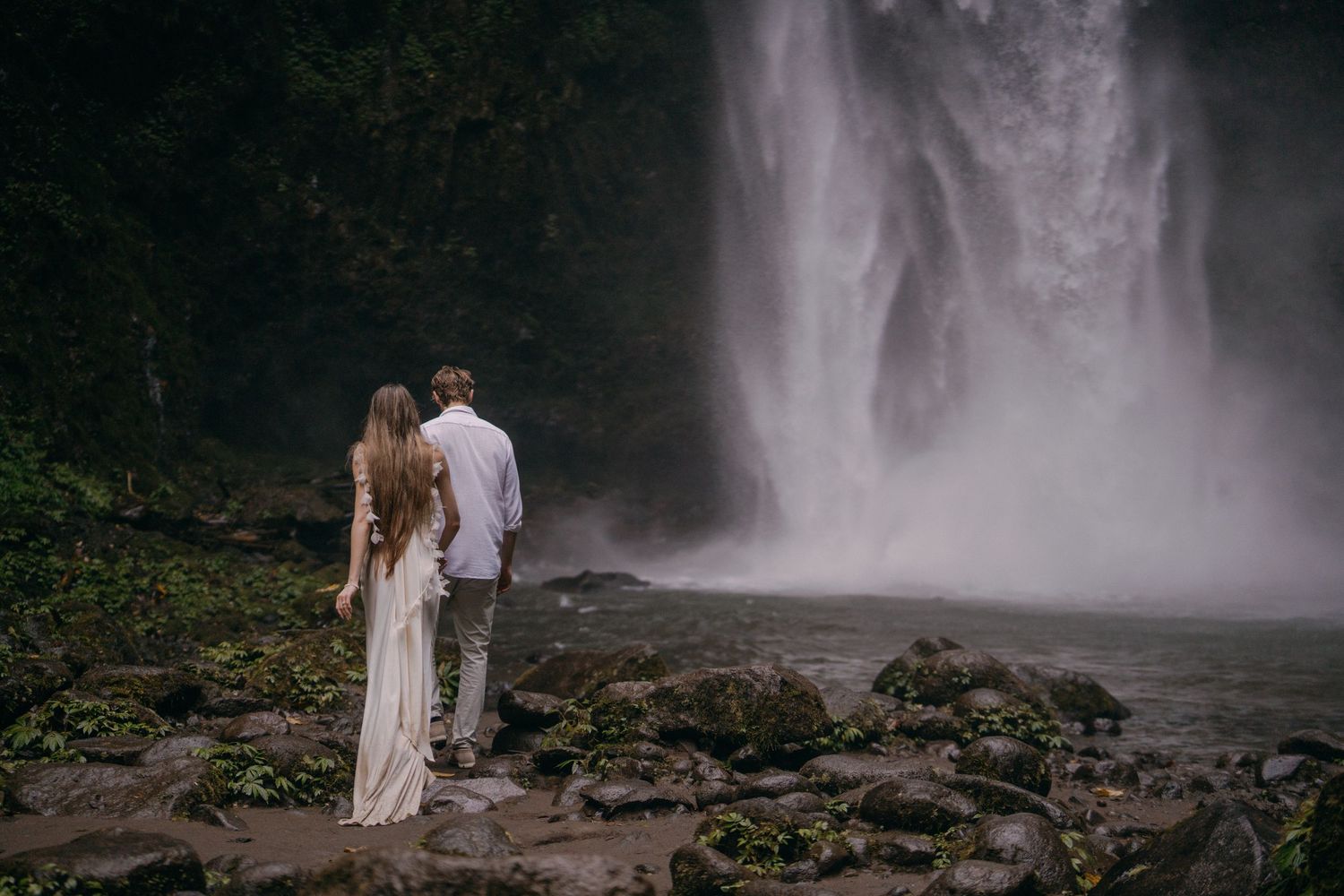 A couple in white outfits stands together near a misty waterfall in a romantic outdoor setting.