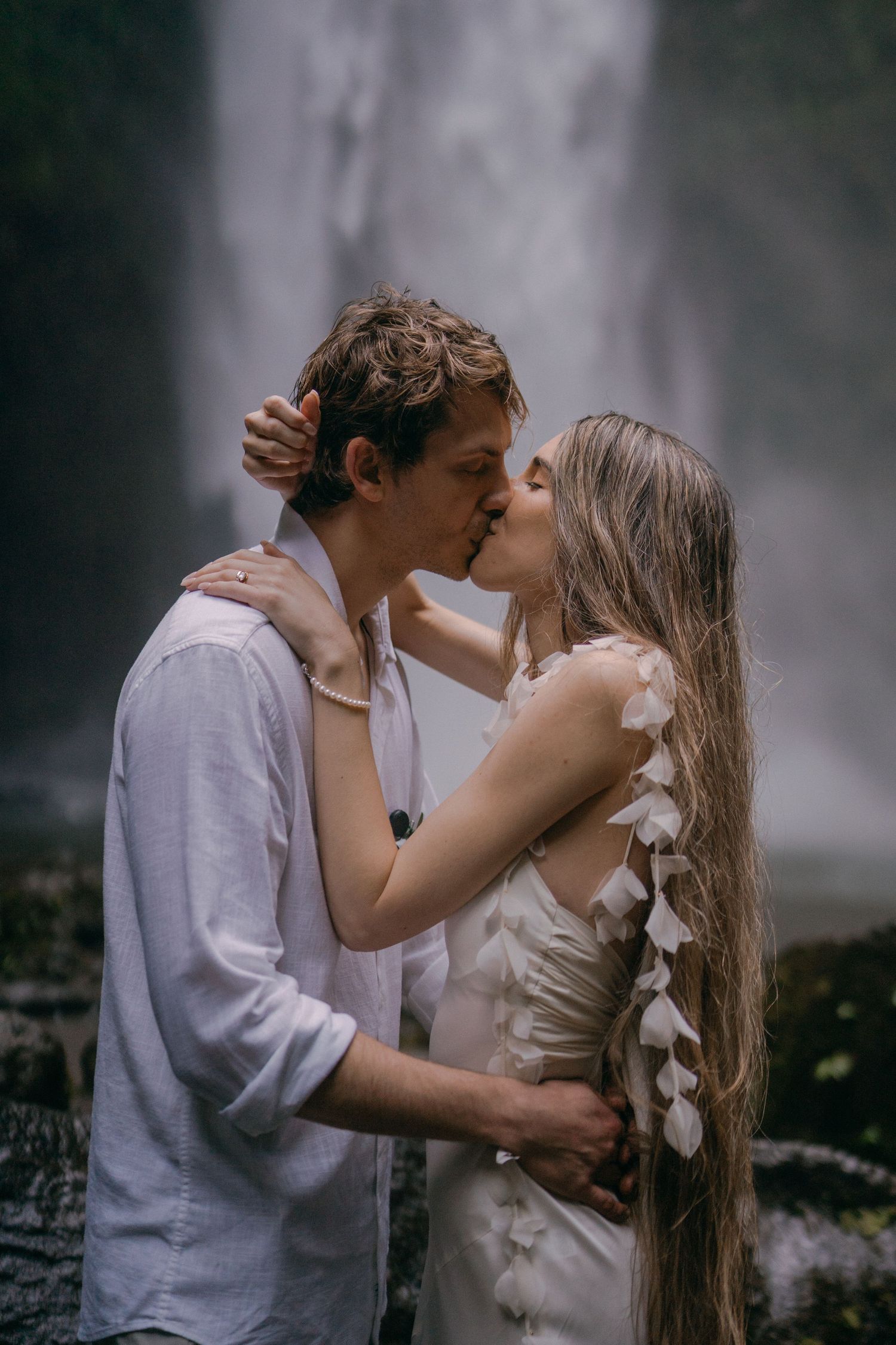 A romantic couple shares a tender kiss in front of a misty waterfall backdrop while wearing light colored clothing.