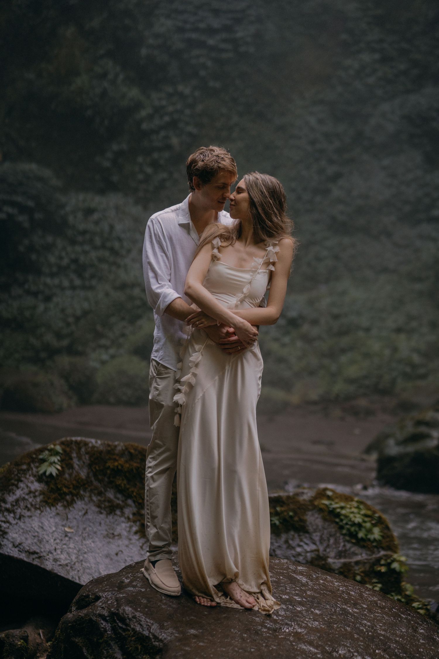 A romantic couple shares an intimate moment on a rocky cliff edge surrounded by moody natural scenery.