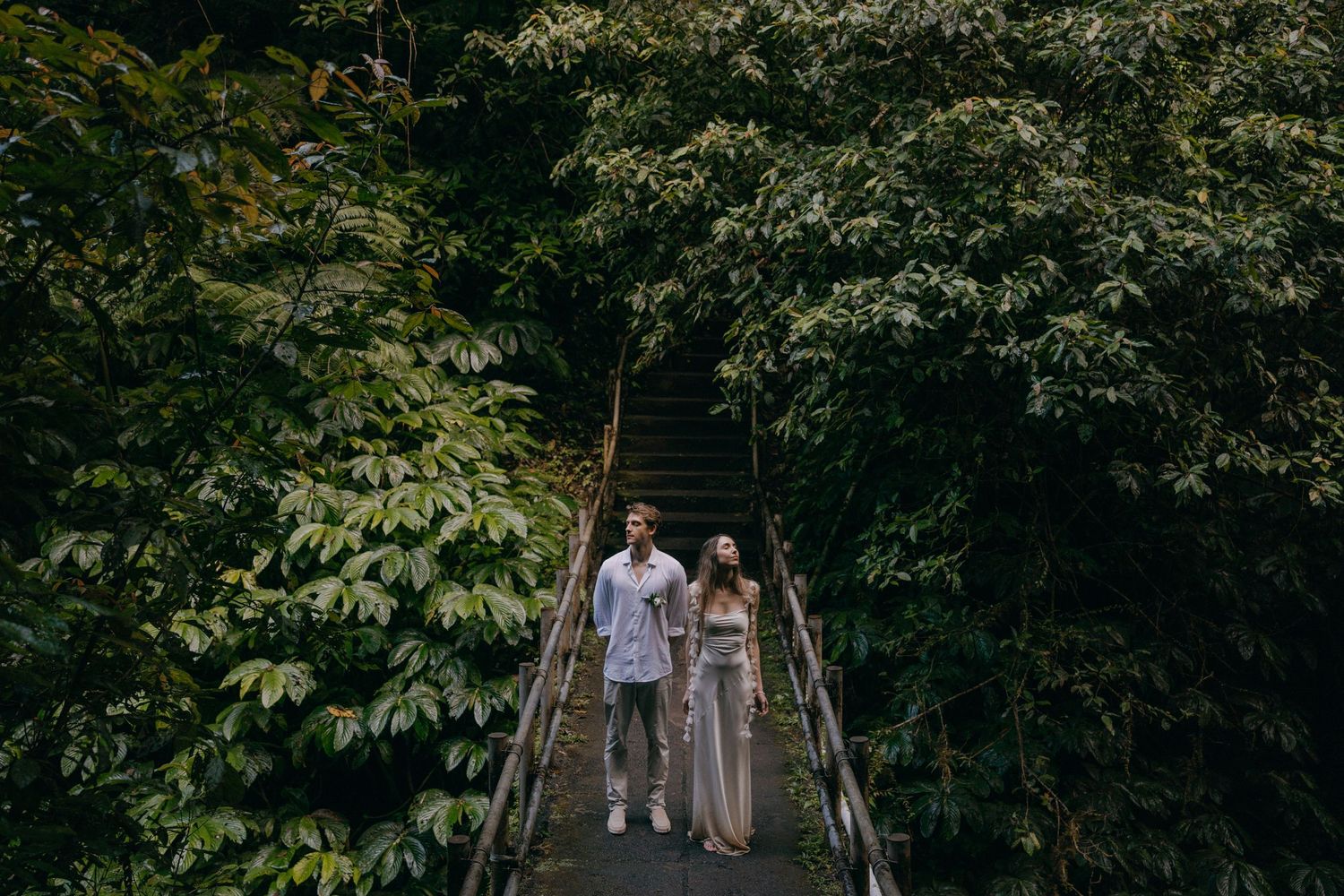 Couple walking together on a wooden bridge surrounded by lush green forest canopy in romantic outdoor setting.