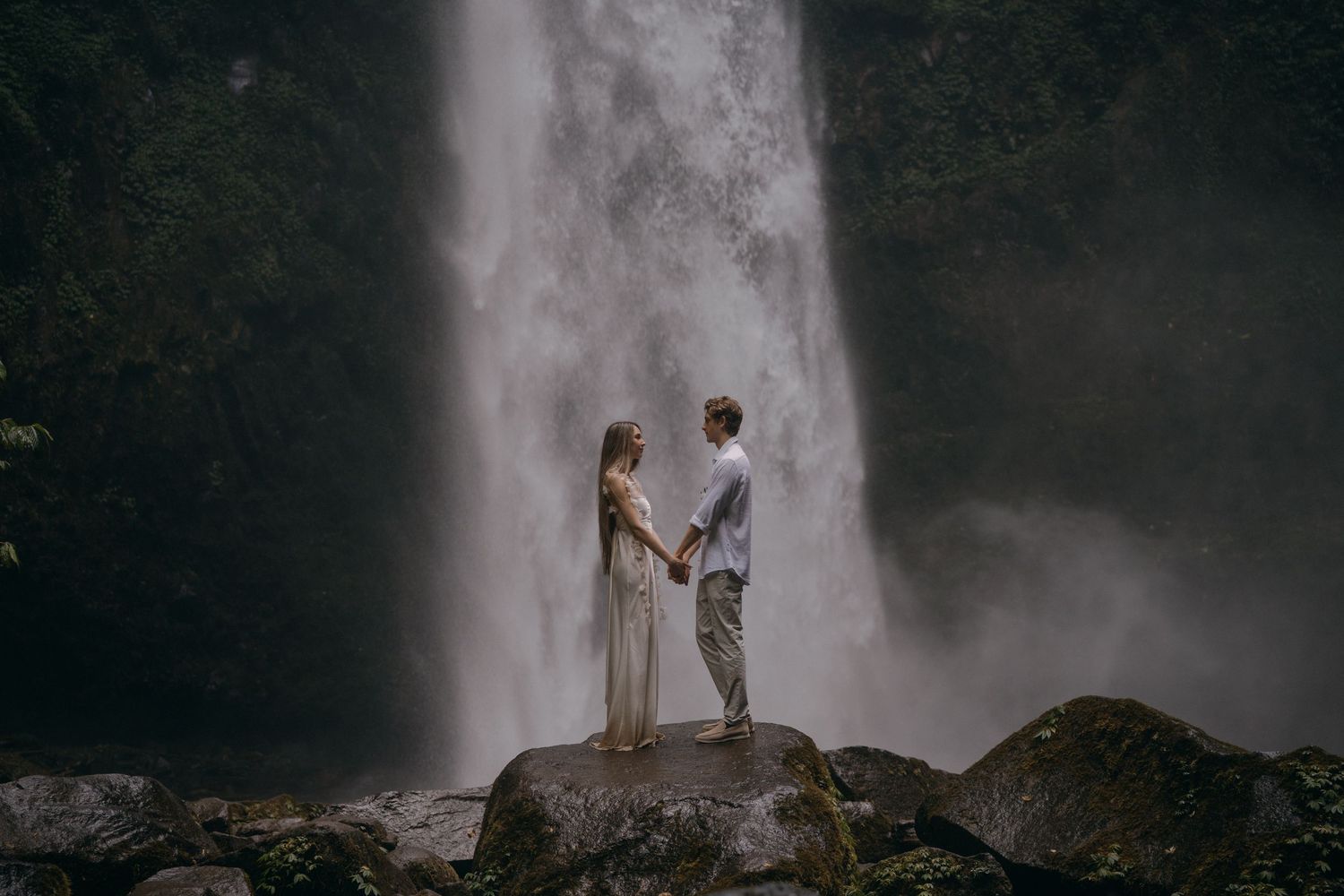 Two people hold hands while standing on rocks in front of a majestic waterfall in a moody, misty setting.