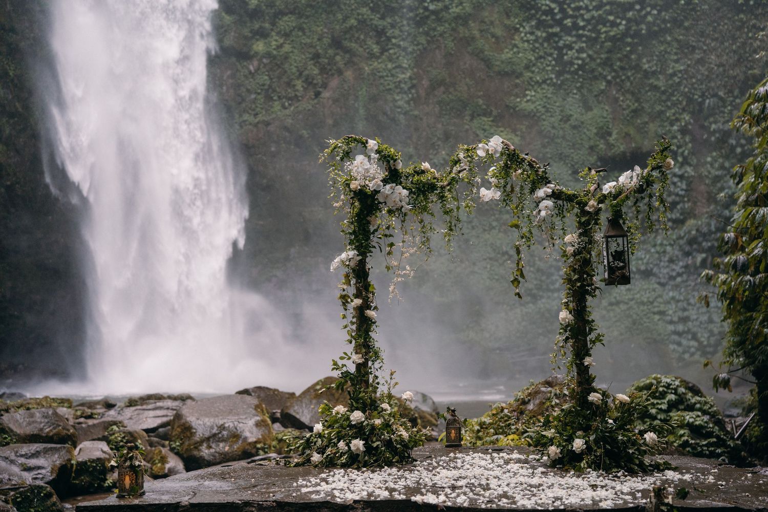 A floral wedding arch decorated with white flowers and greenery stands before a misty waterfall in nature.