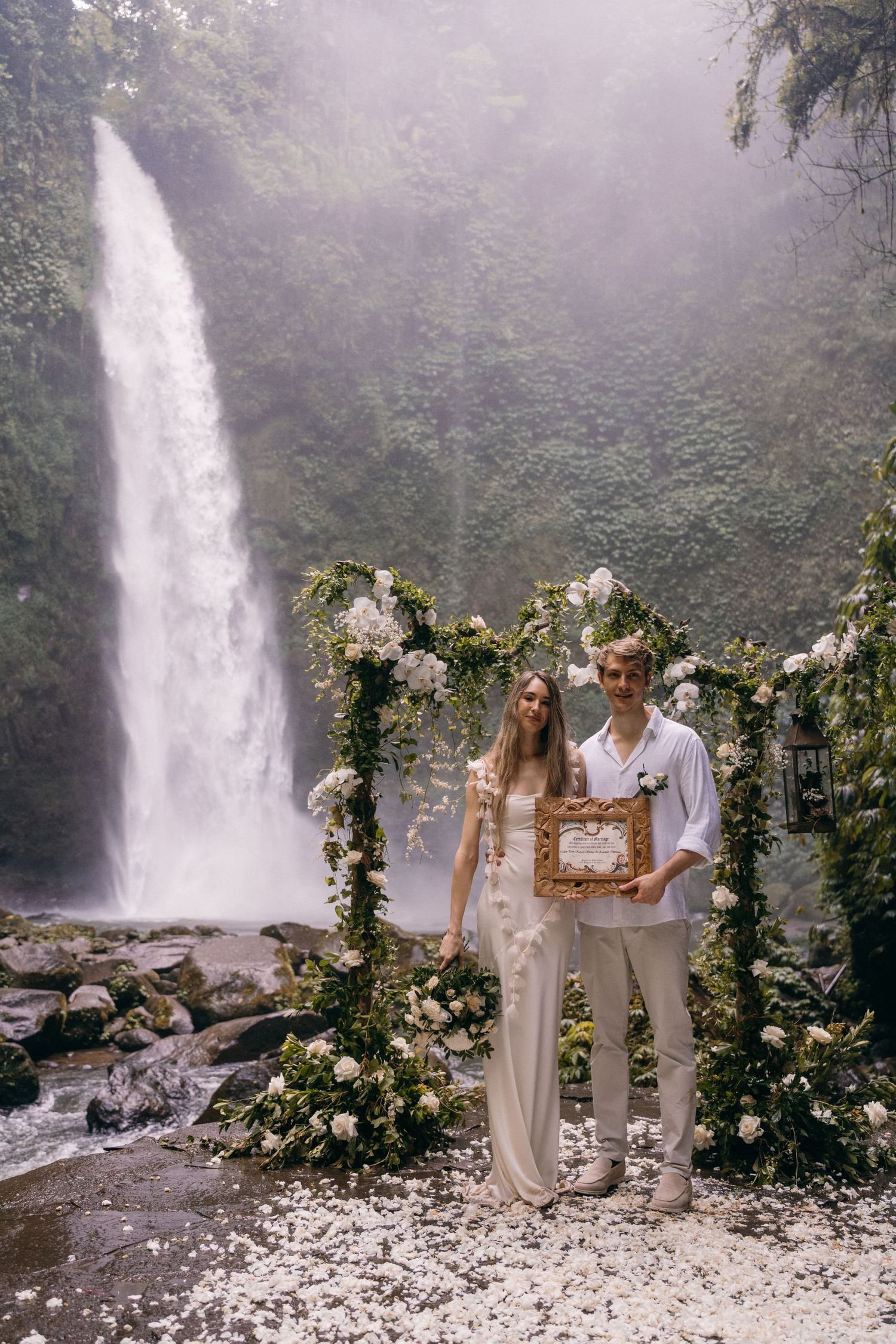 A floral arch adorns a scenic waterfall setting with mist and lush greenery in the background.