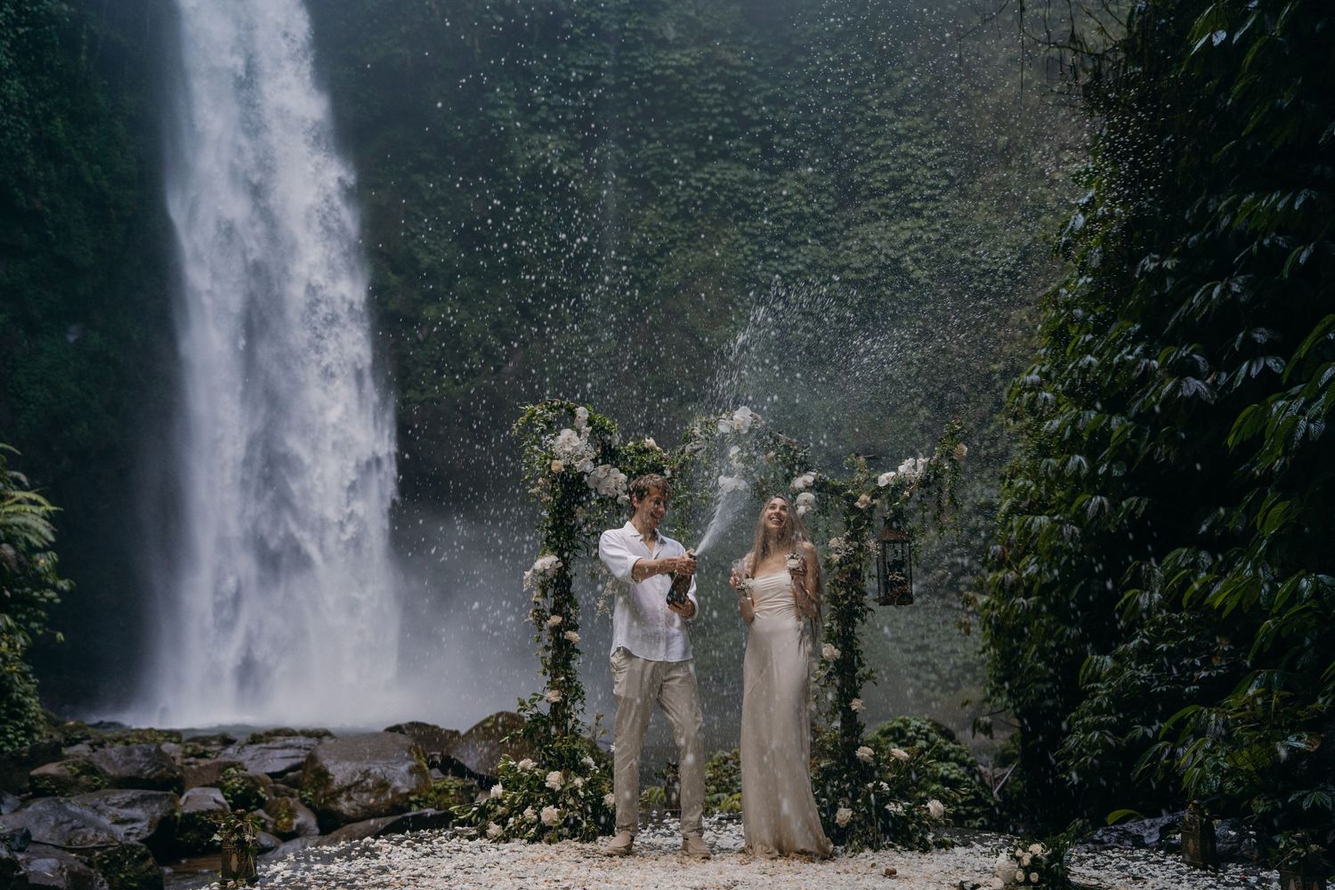 People celebrating near a majestic waterfall in a lush tropical setting as mist rises in the background.