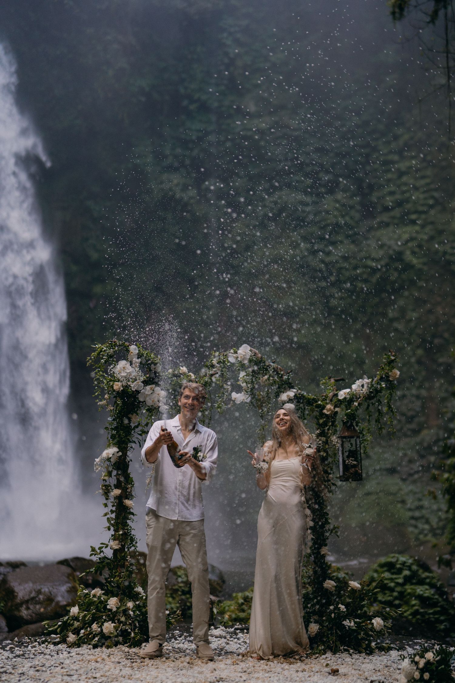 Couple celebrates with confetti and flower petals in front of a majestic waterfall during an outdoor wedding ceremony.