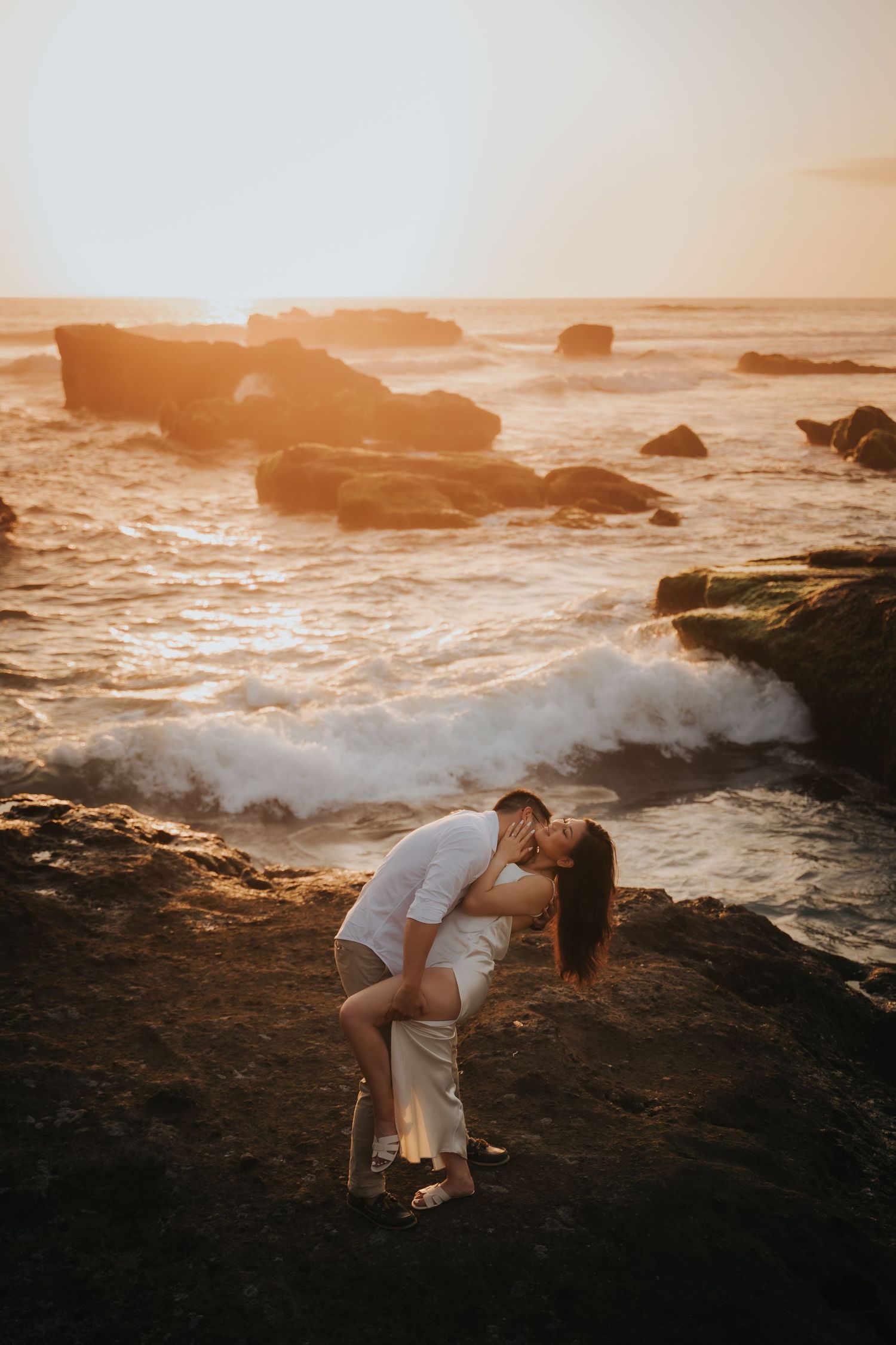 Couple sharing a romantic dip kiss on rocky coastline at golden sunset with crashing ocean waves behind them.