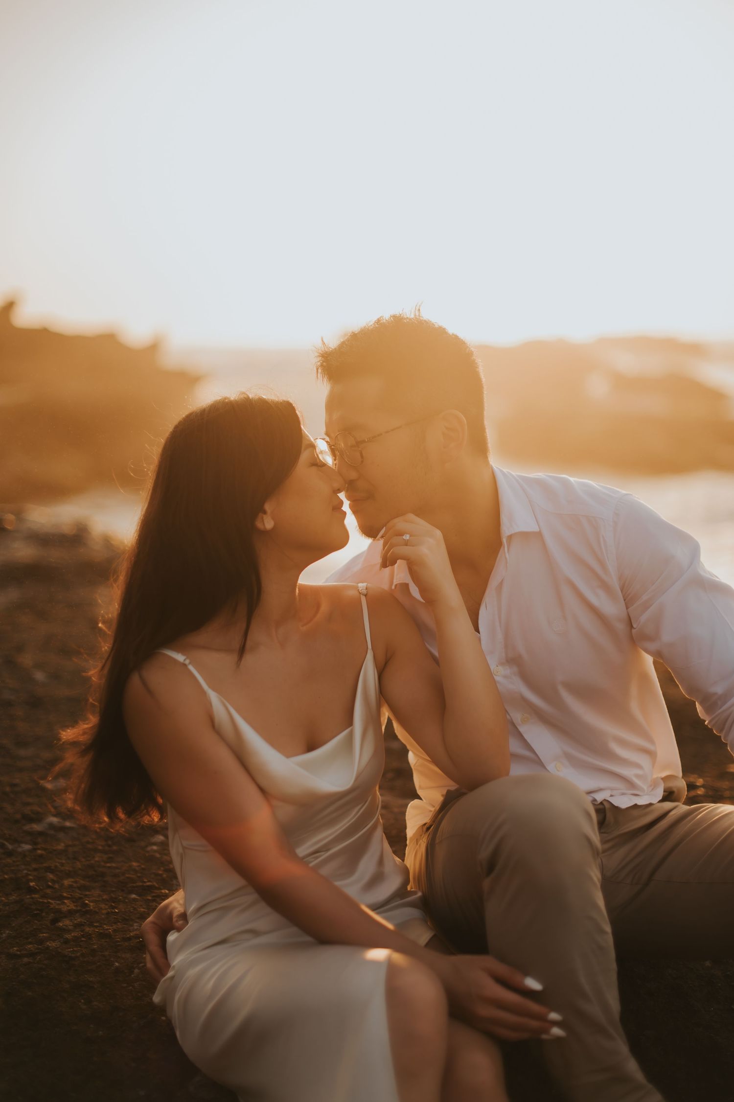 Couple sharing a romantic moment at golden hour sunset, woman in white dress and man in white shirt sitting outdoors.