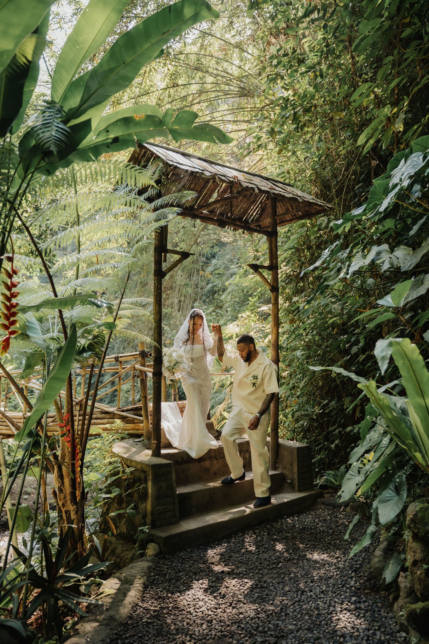 Bride and groom pose under rustic thatched gazebo surrounded by lush tropical jungle greenery and foliage.