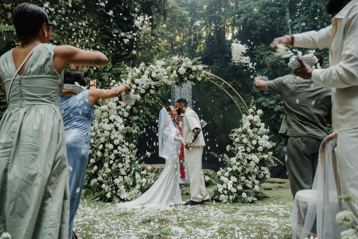 Couple shares a kiss under a white floral arch as guests toss petals at outdoor wedding.
