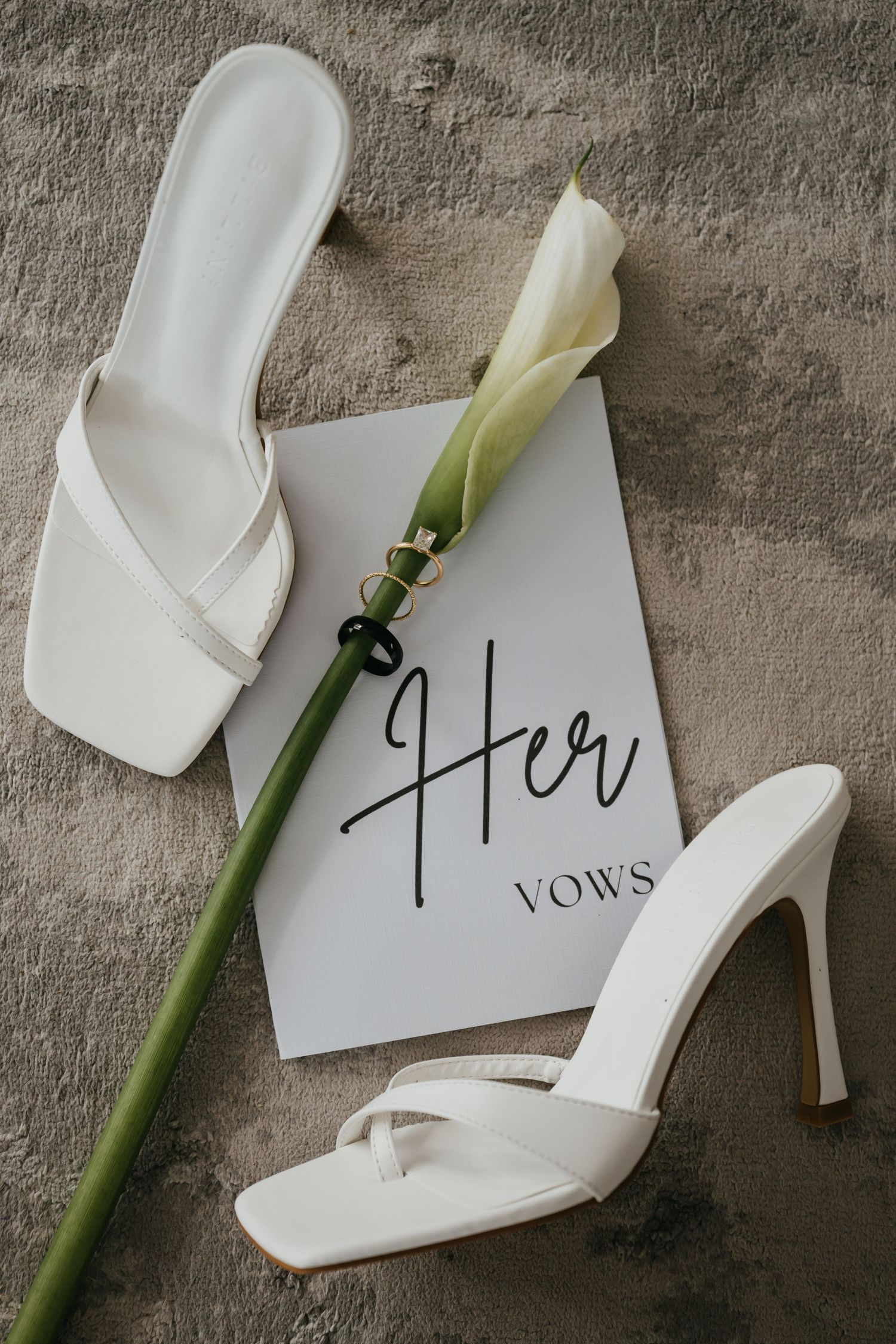 White bridal heels and vow book with calla lily and wedding rings on grey textured surface.
