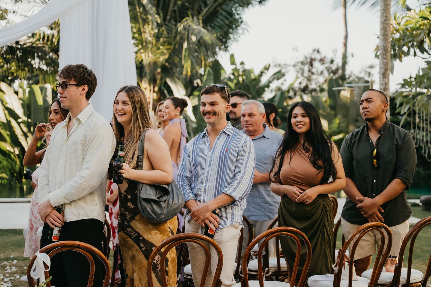 Smiling wedding guests standing and seated at an outdoor tropical ceremony with palm trees in the background.