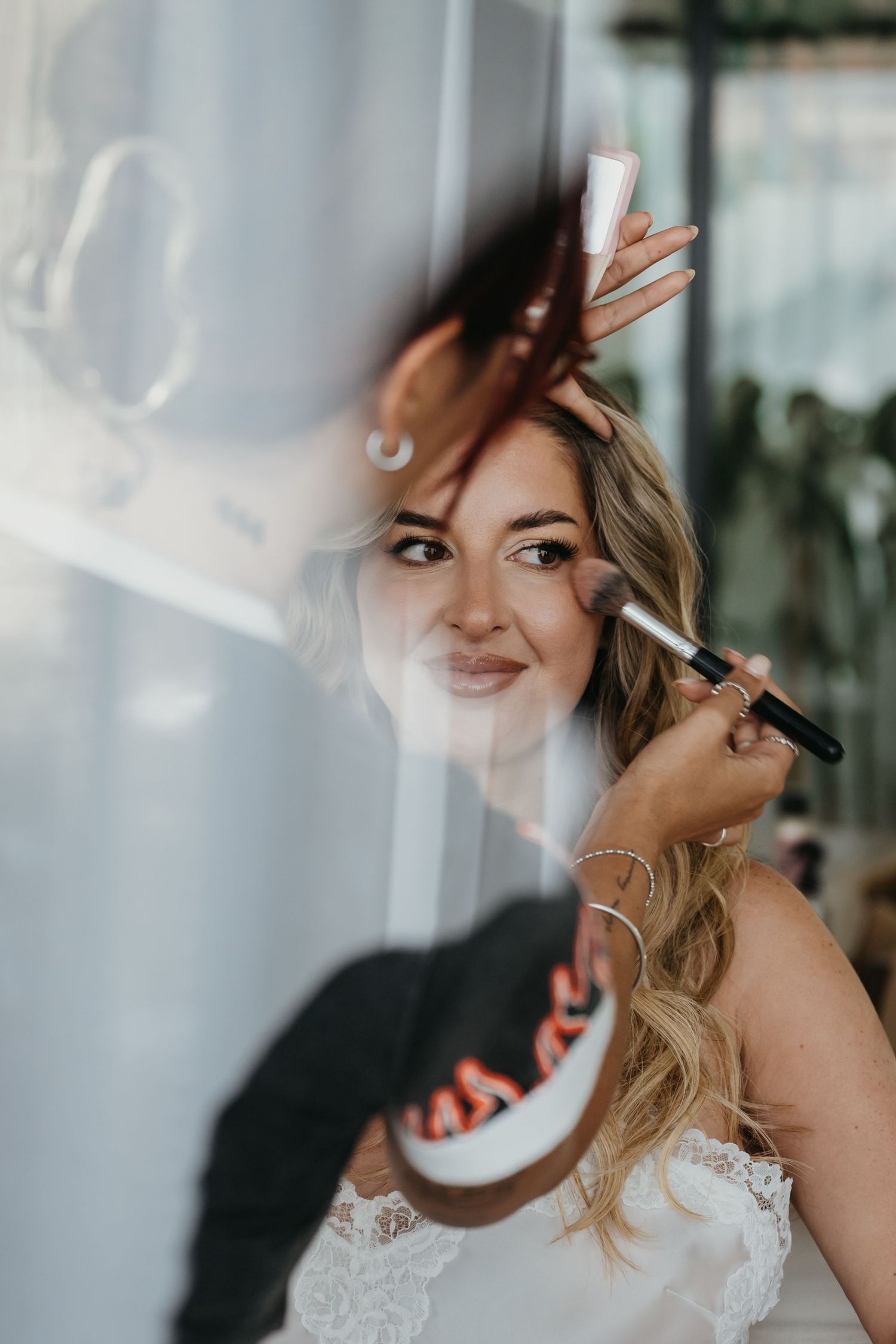 Bride getting makeup applied with brush, smiling at reflection in mirror, wearing white lace dress.
