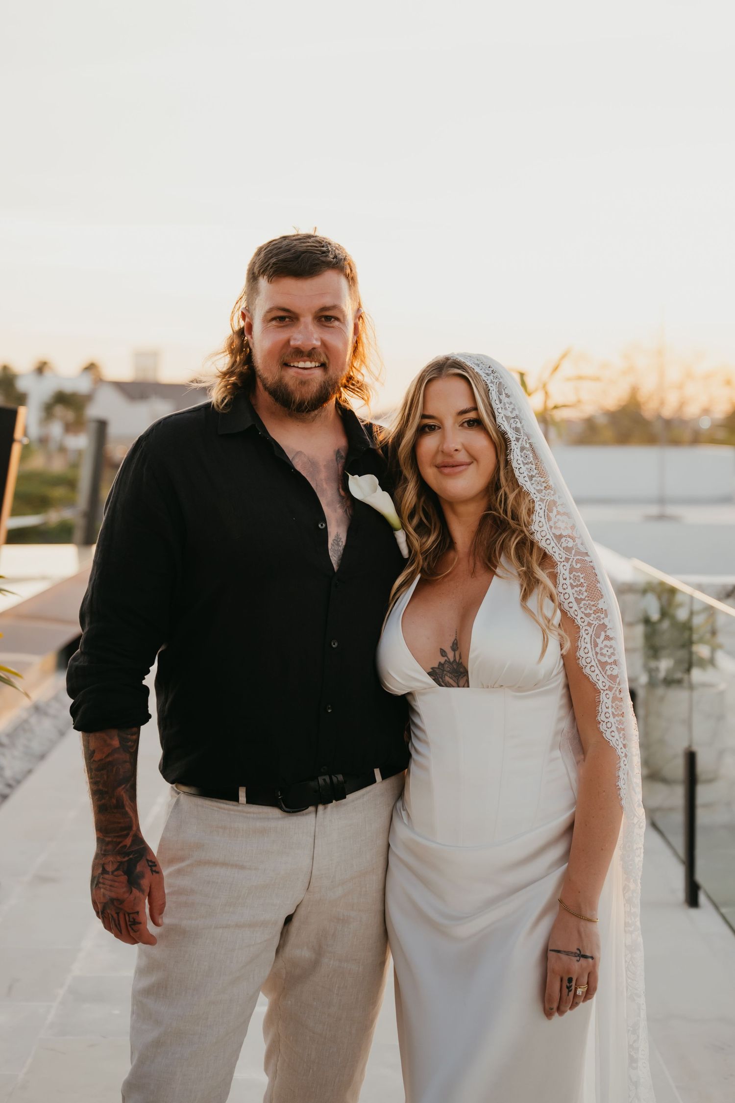 Tattooed couple posing together at sunset rooftop, groom in black shirt and bride in white gown.