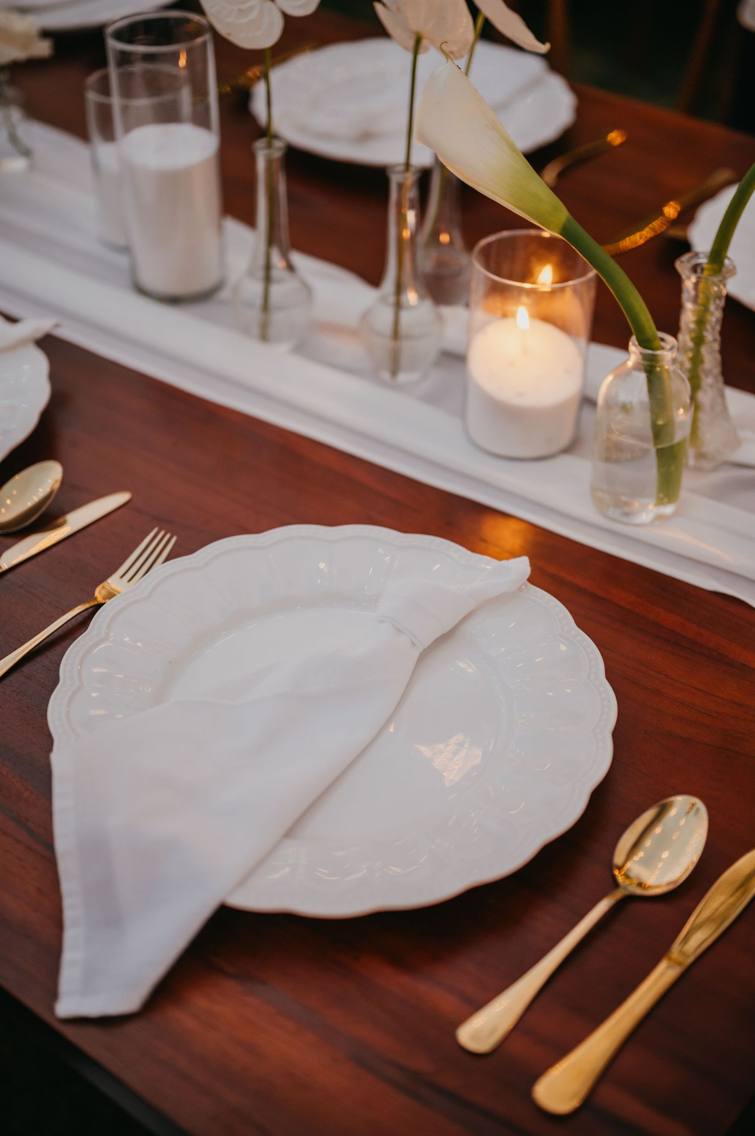 Elegant wedding table setting with white embossed plate, gold cutlery, lit candle, and white calla lily.