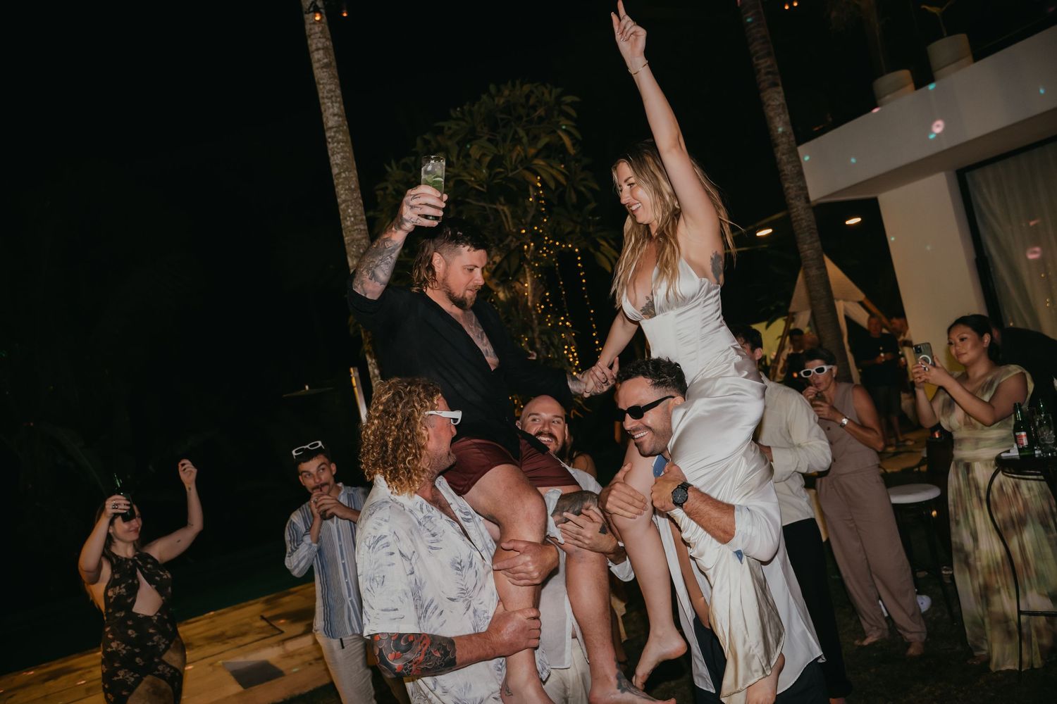 Bride and groom are lifted by guests in a lively crowd celebration during their outdoor wedding reception.