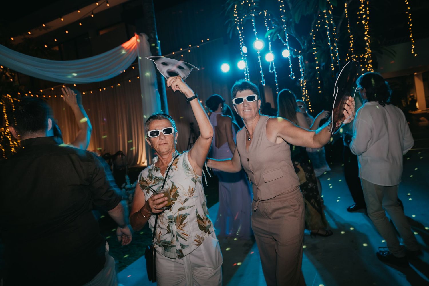 Two wedding guests dance energetically wearing novelty sunglasses on lit reception dance floor.