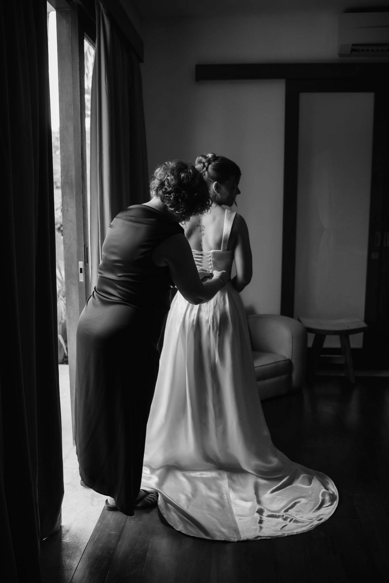 Black and white photo of bridesmaid helping bride with wedding dress near window.