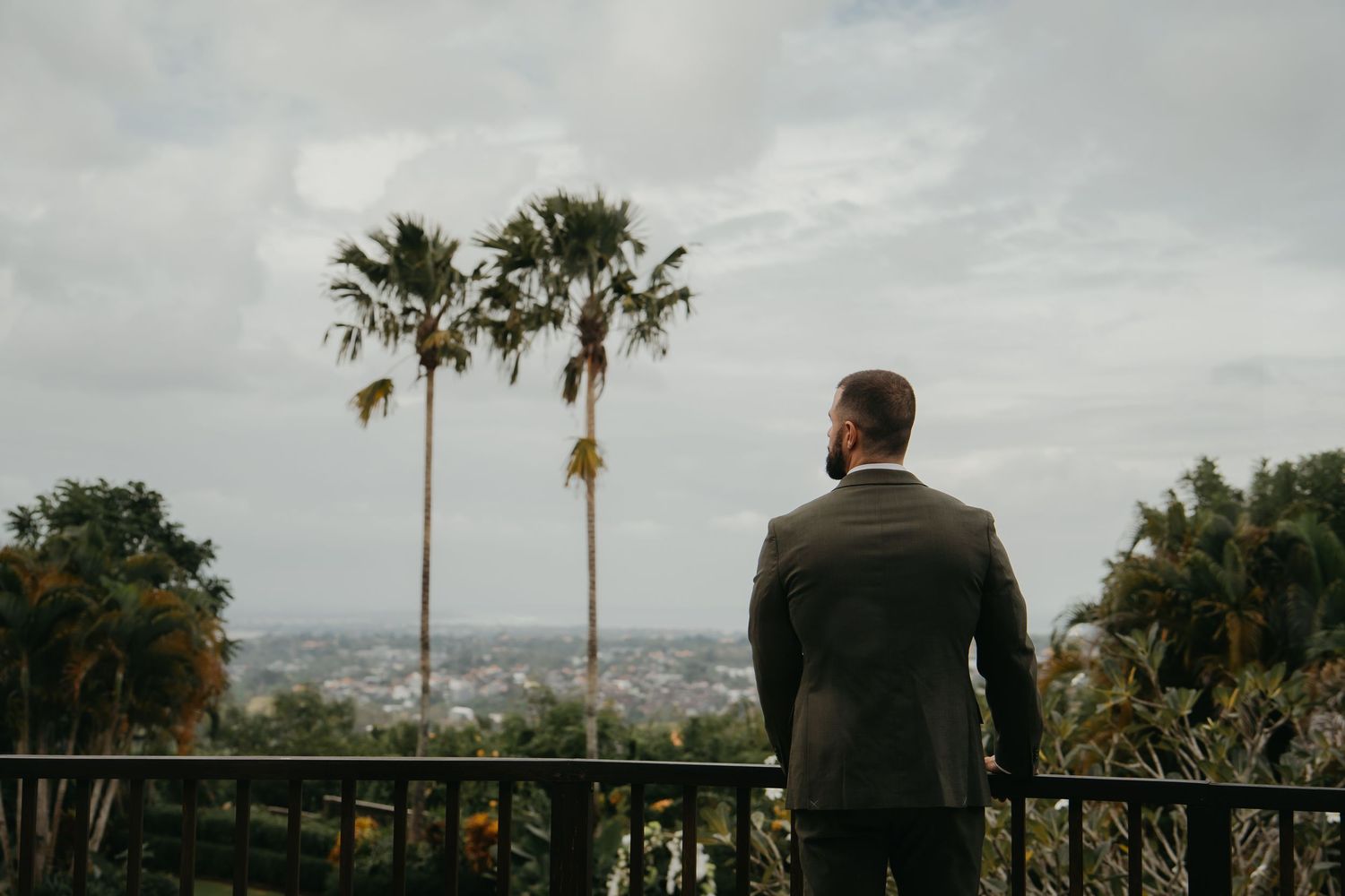 Groom in grey suit standing on balcony overlooking tropical cityscape with palm trees.
