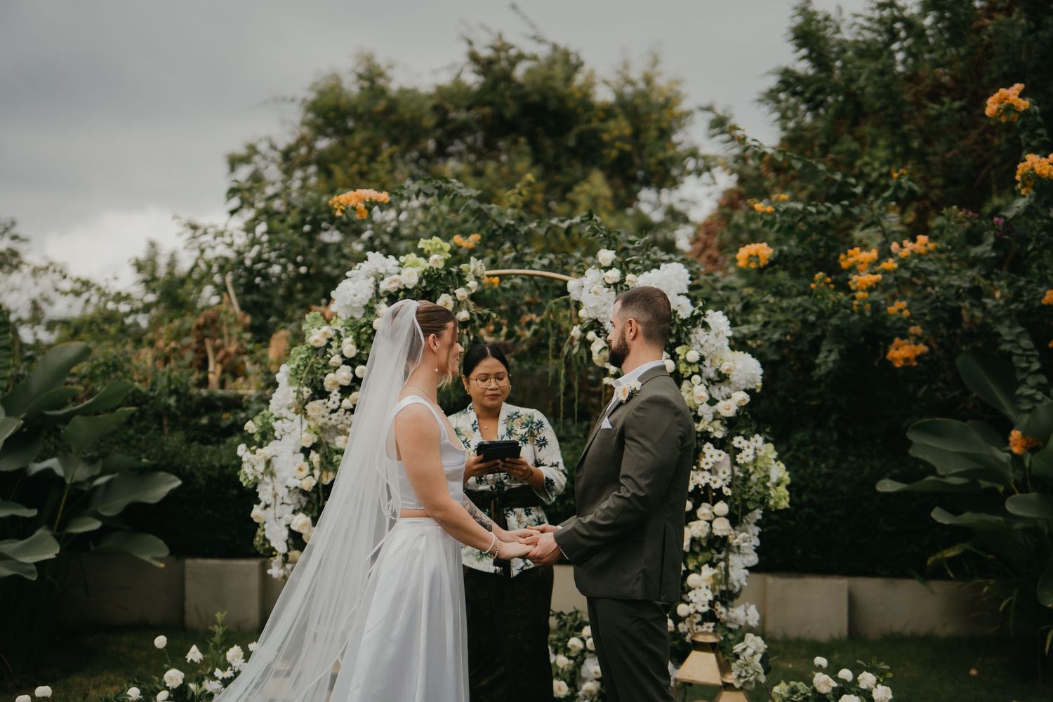 Bride and groom exchange vows under white floral arch at outdoor garden wedding ceremony with officiant.