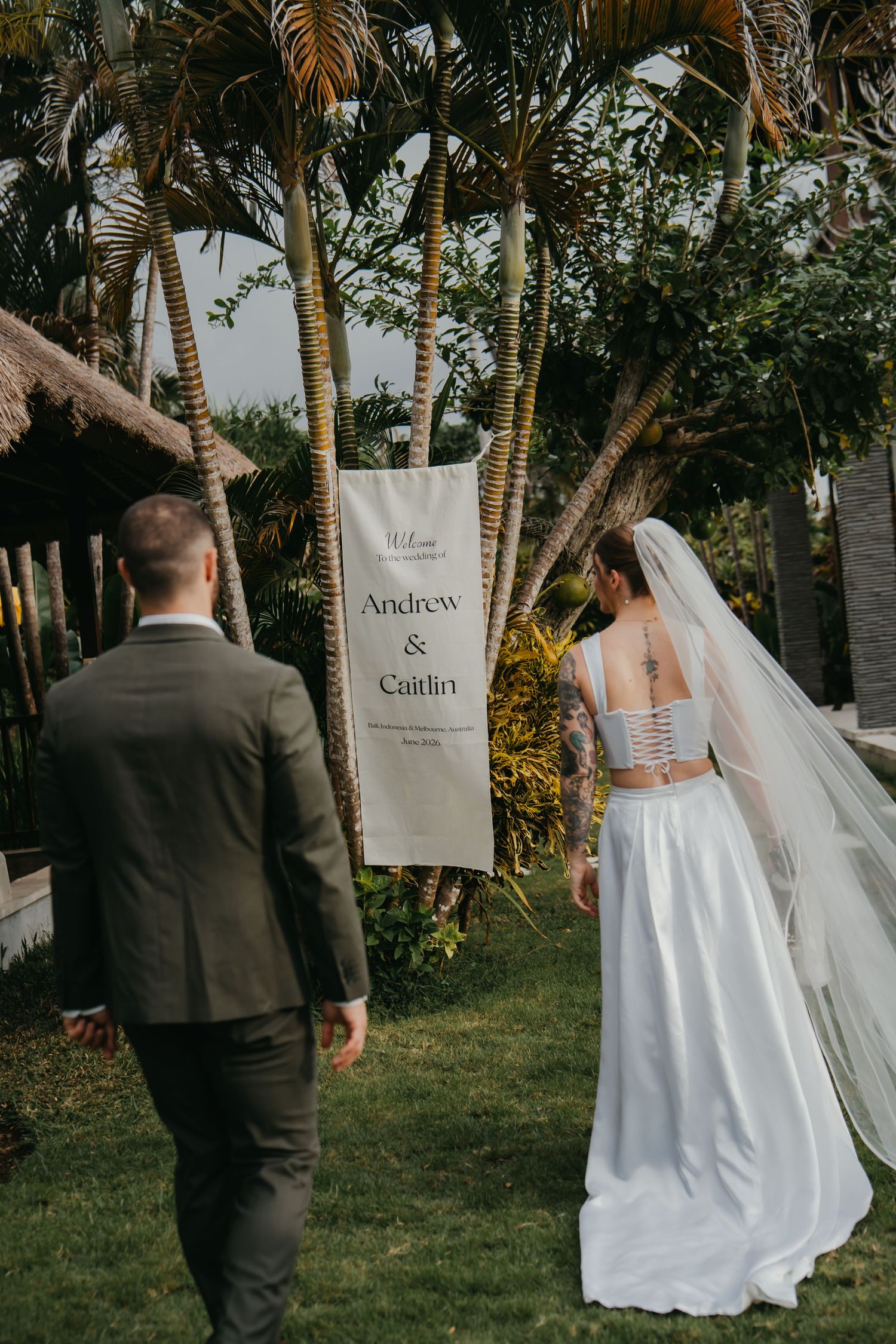 Bride and groom walk past welcome banner reading Andrew and Caitlin at tropical outdoor wedding venue.