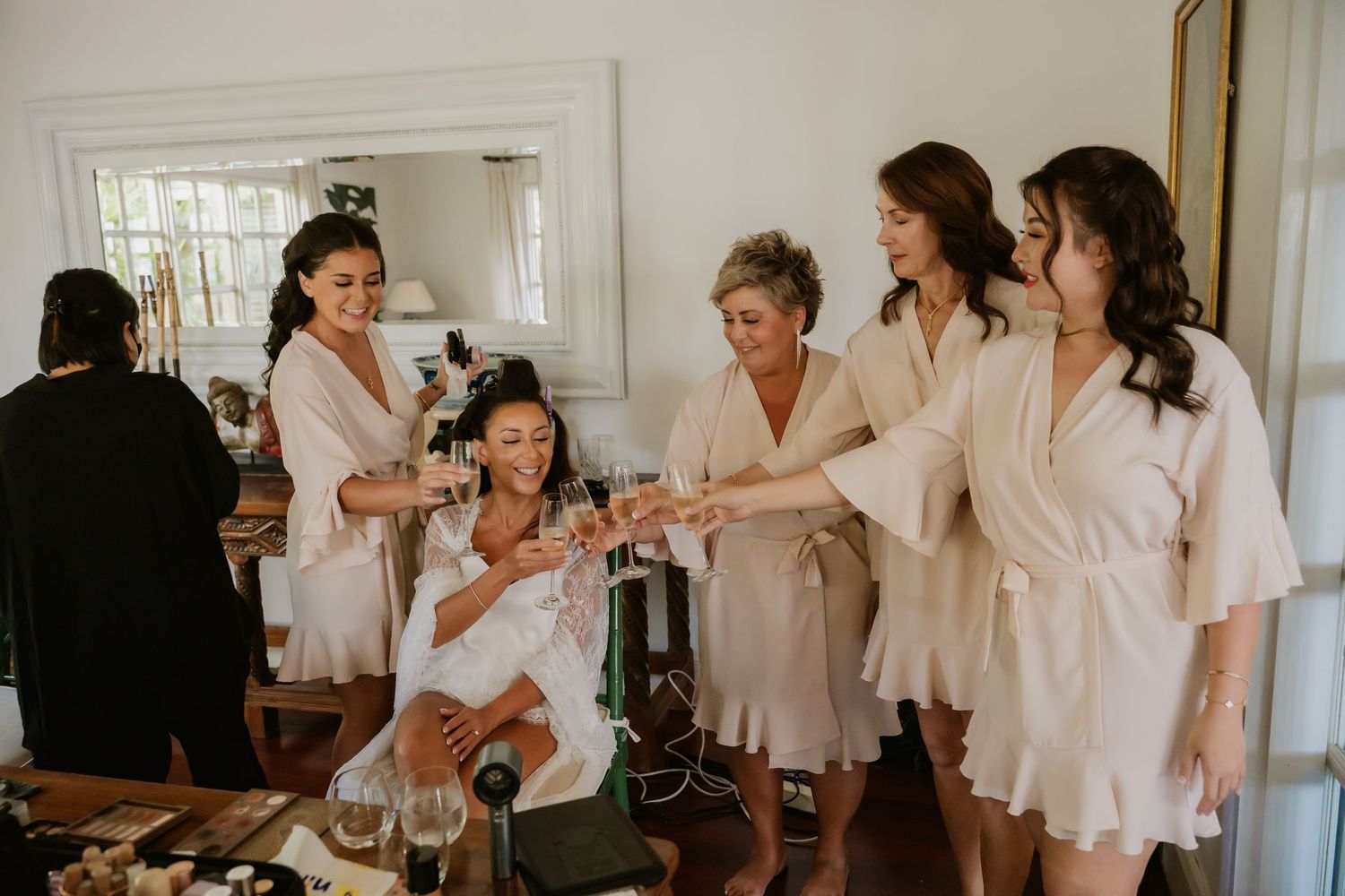 Bridesmaids in white robes toast with the bride during wedding morning preparations indoors.