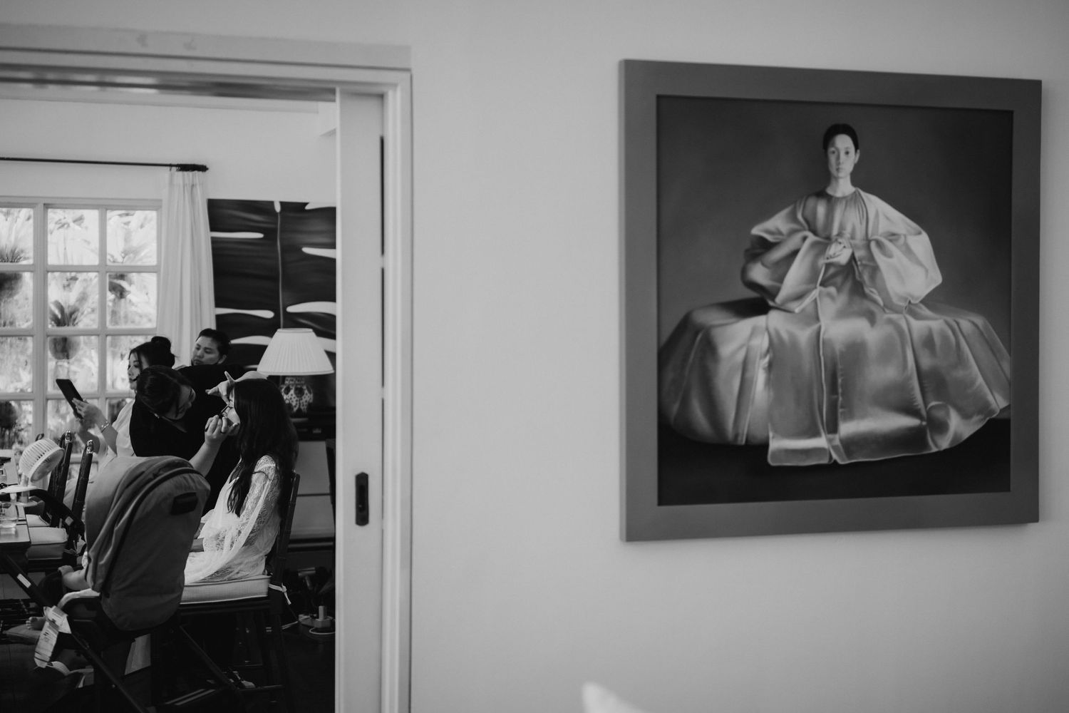 Black and white photo of a framed portrait on a wall with bridesmaids getting ready in a room behind it.