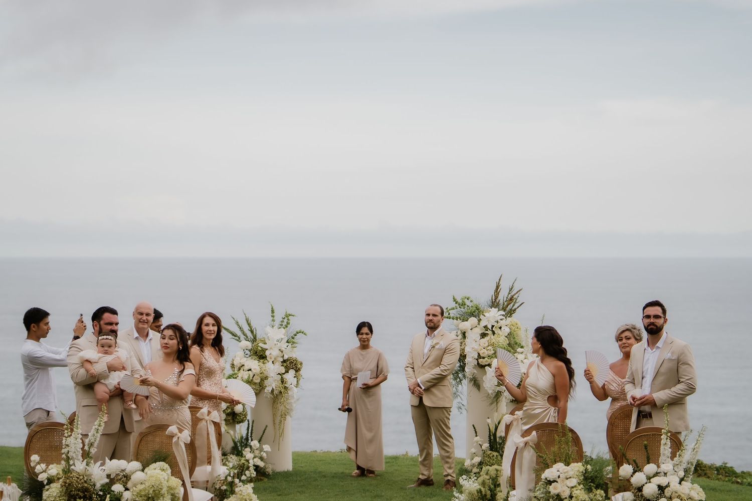 Wedding party stands at floral altar with ocean backdrop on lush green lawn.