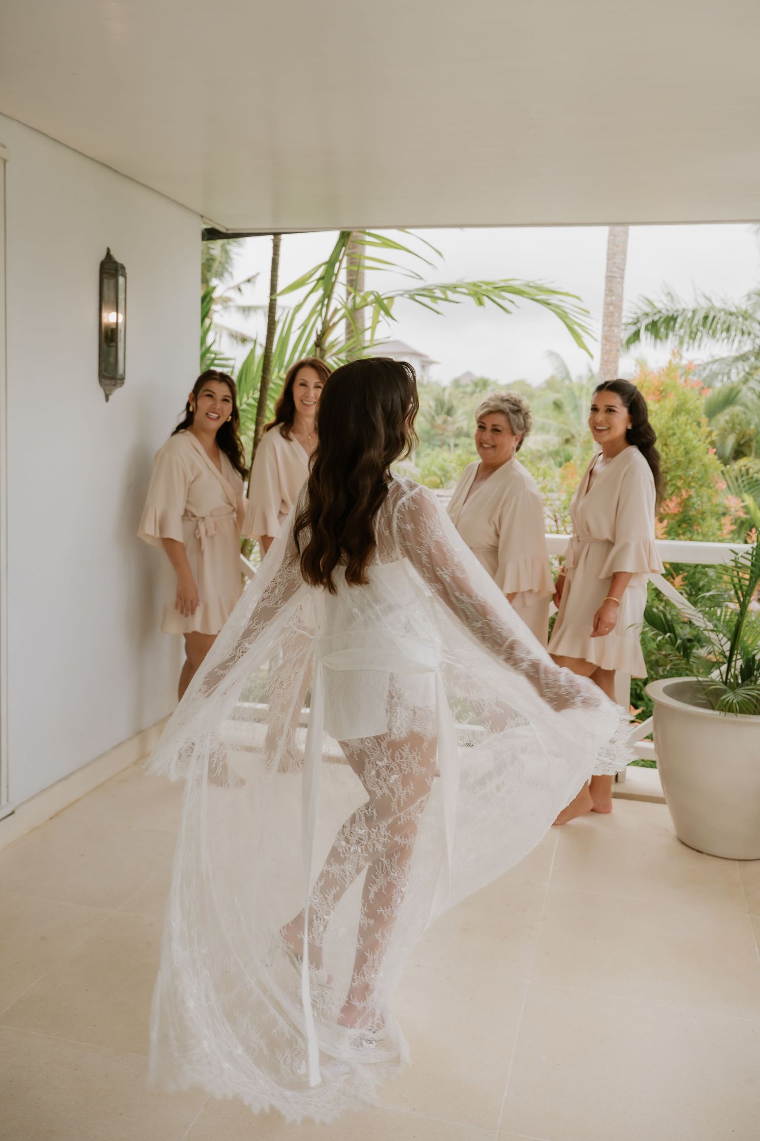 Bride in lace robe twirls before bridesmaids in matching robes in a tropical villa getting ready room.