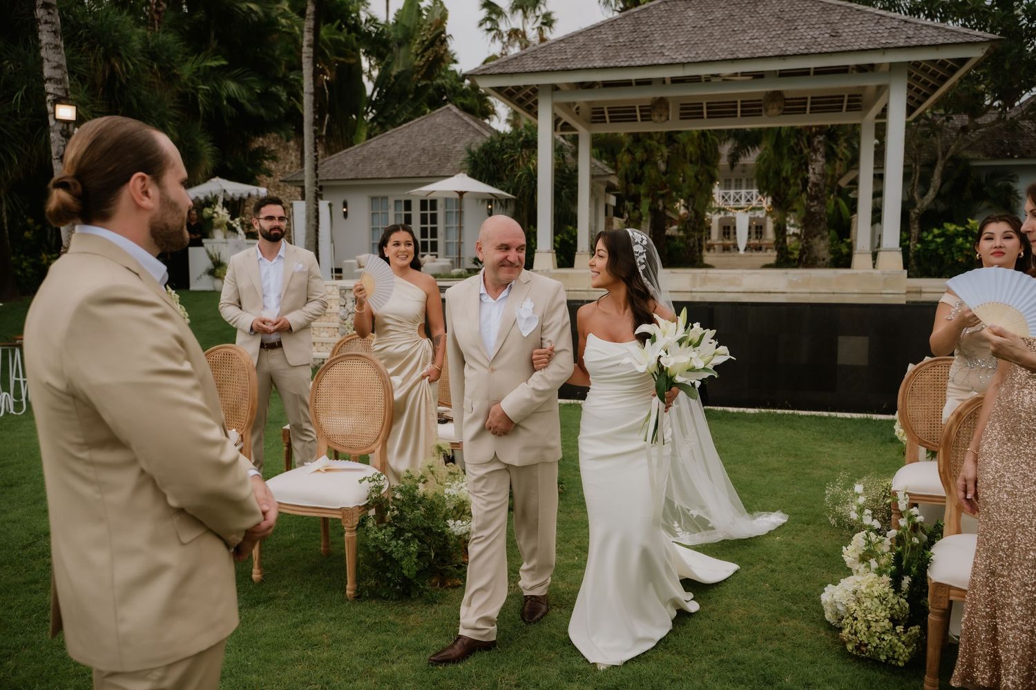 Bride in a fitted white gown being walked down the aisle by her father at a lush tropical garden wedding.