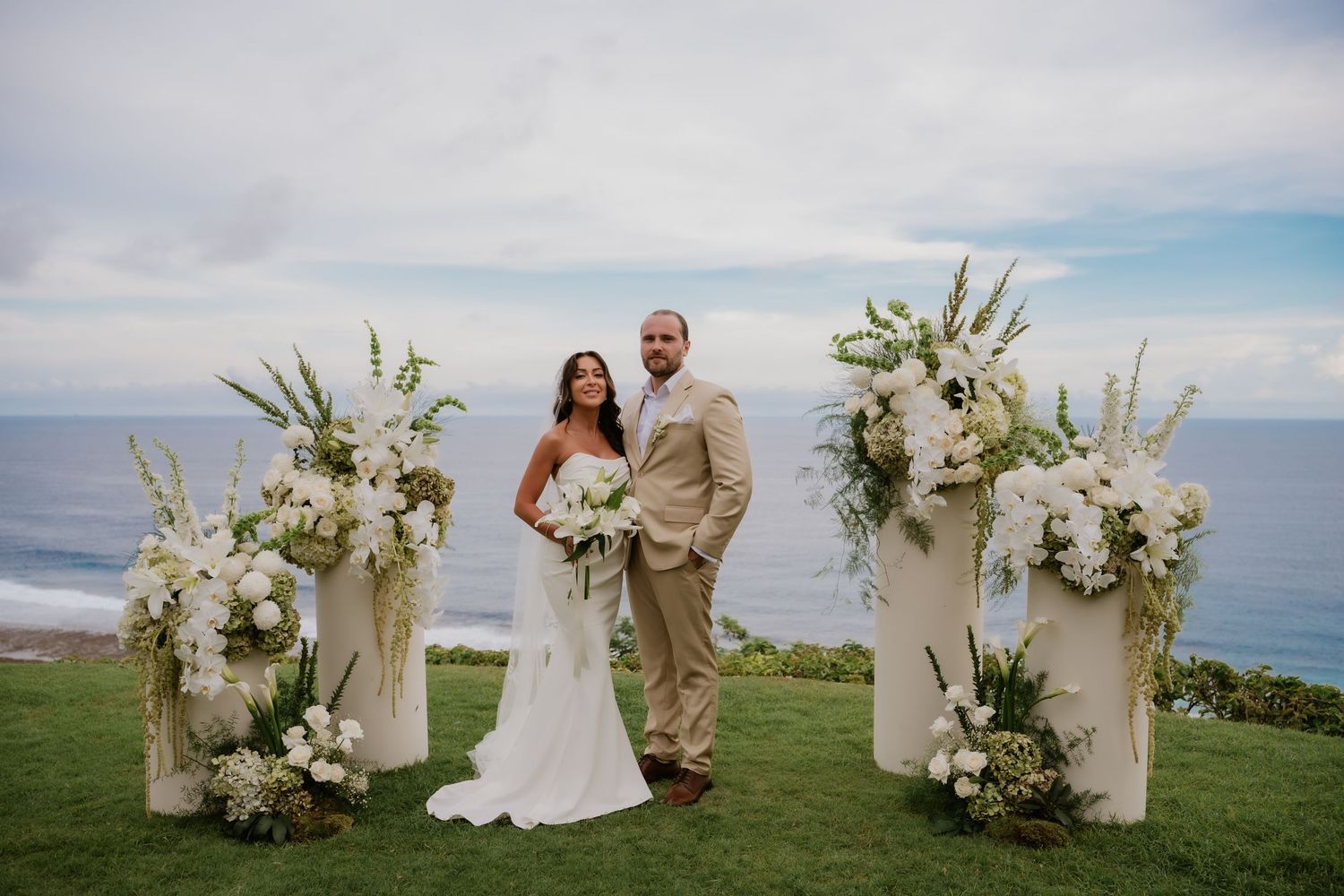 Bride and groom pose between white pillar floral arrangements with ocean view at outdoor wedding.