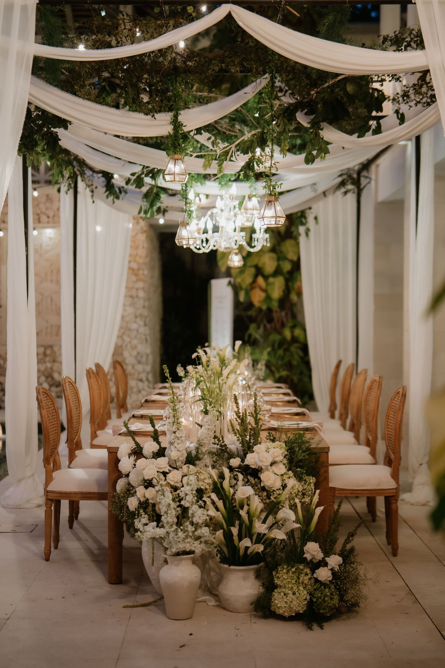 Romantic outdoor wedding dinner table adorned with white flowers, greenery, and draped fabric ceiling.