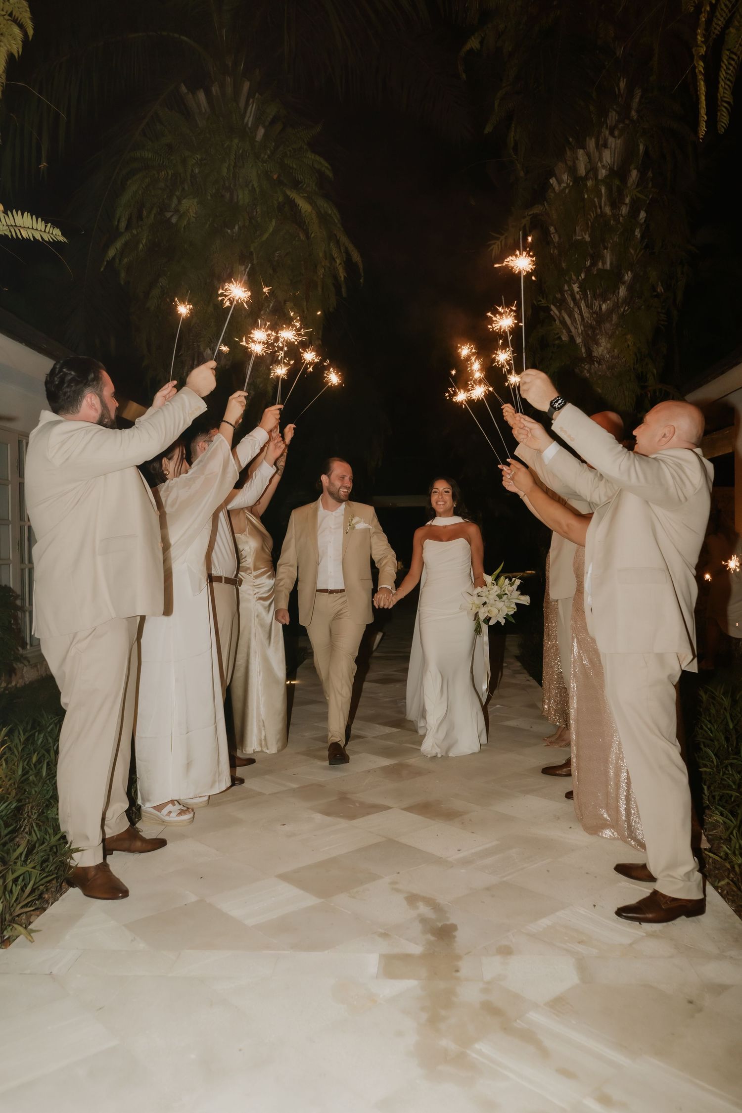 Newlyweds walking through a sparkler tunnel with wedding party celebrating at night under palm trees.