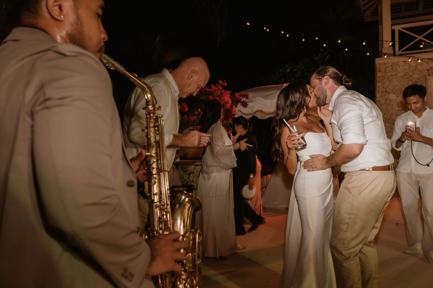 Bride and groom kiss on dance floor while saxophonist performs at outdoor wedding reception.