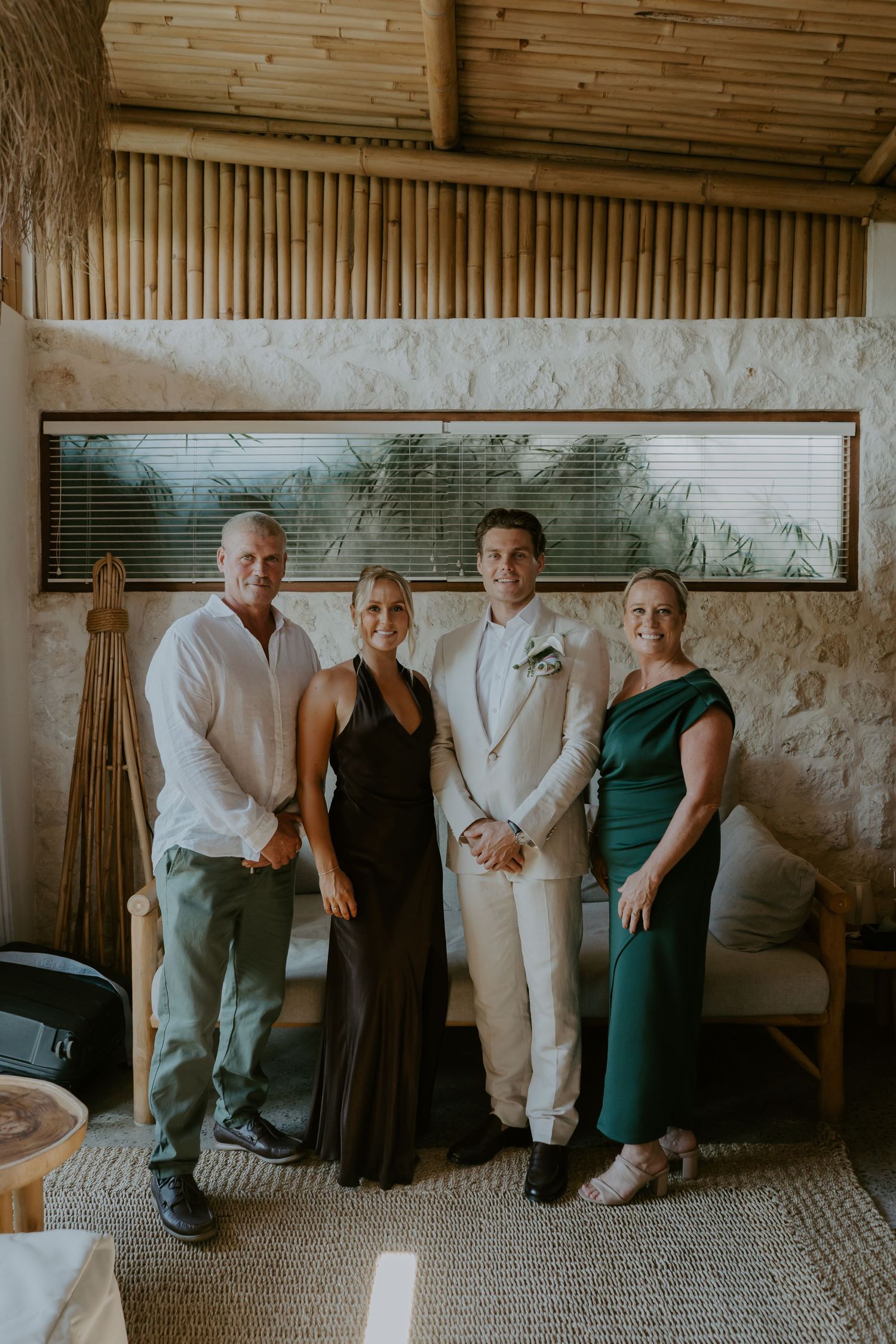 Groom in white suit poses with family indoors at tropical bamboo wedding venue.