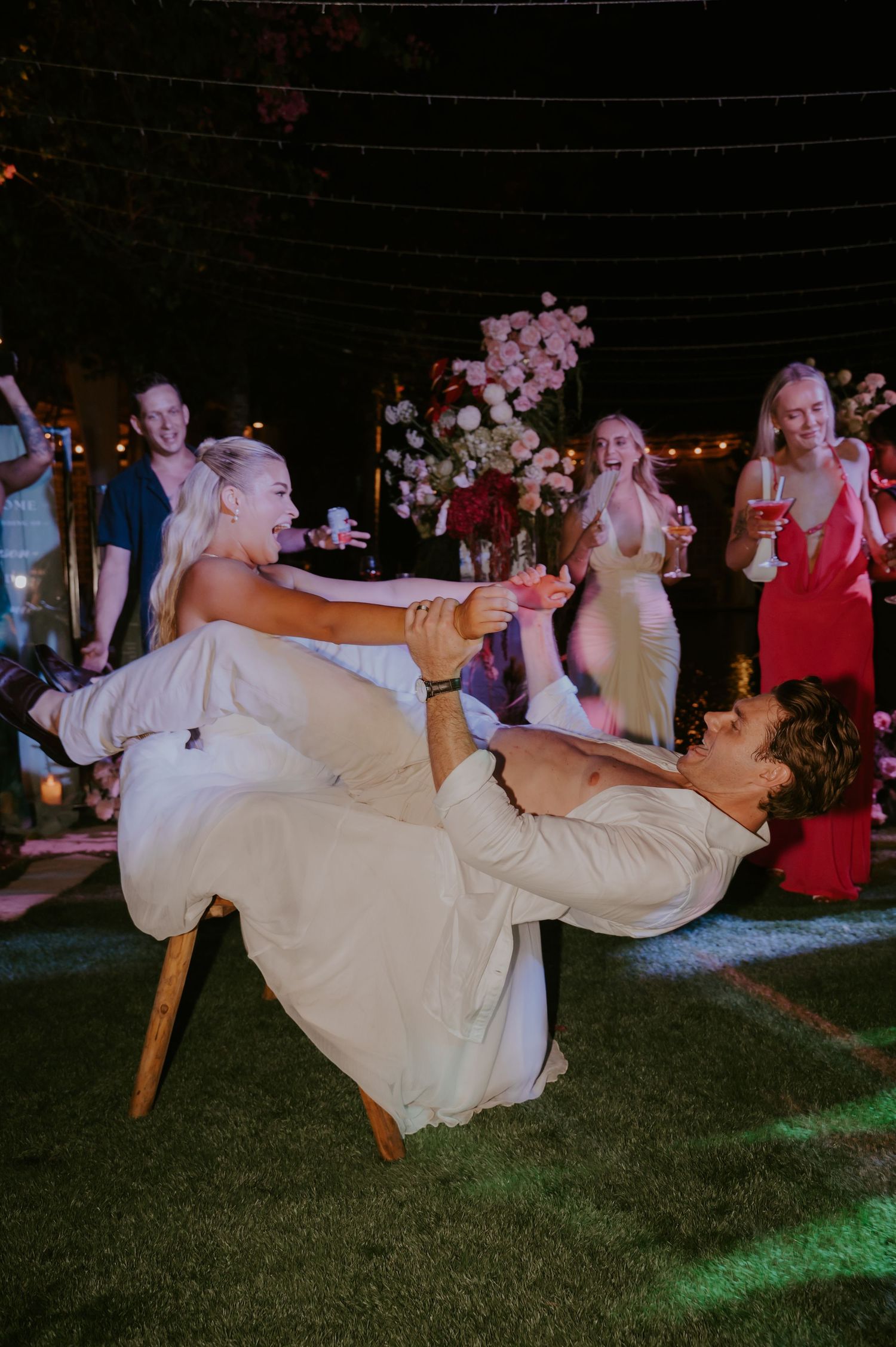 Groom balancing bride on chair during fun wedding reception game on grass dance floor.