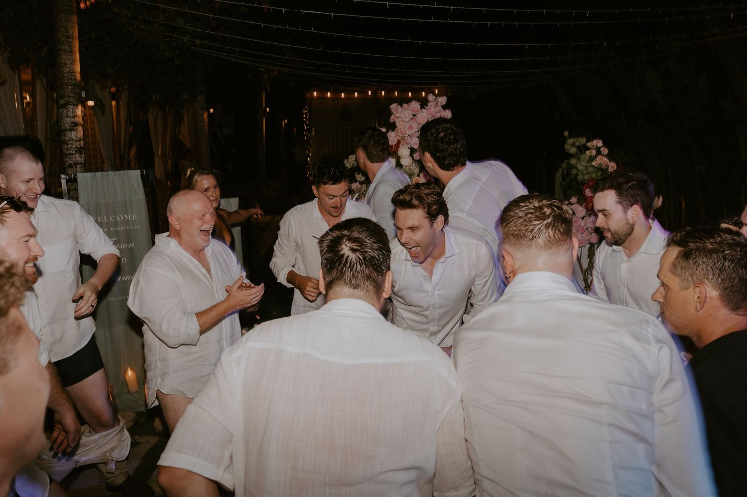 Groomsmen in white shirts huddle together in a lively group celebration on the dance floor at a wedding reception.